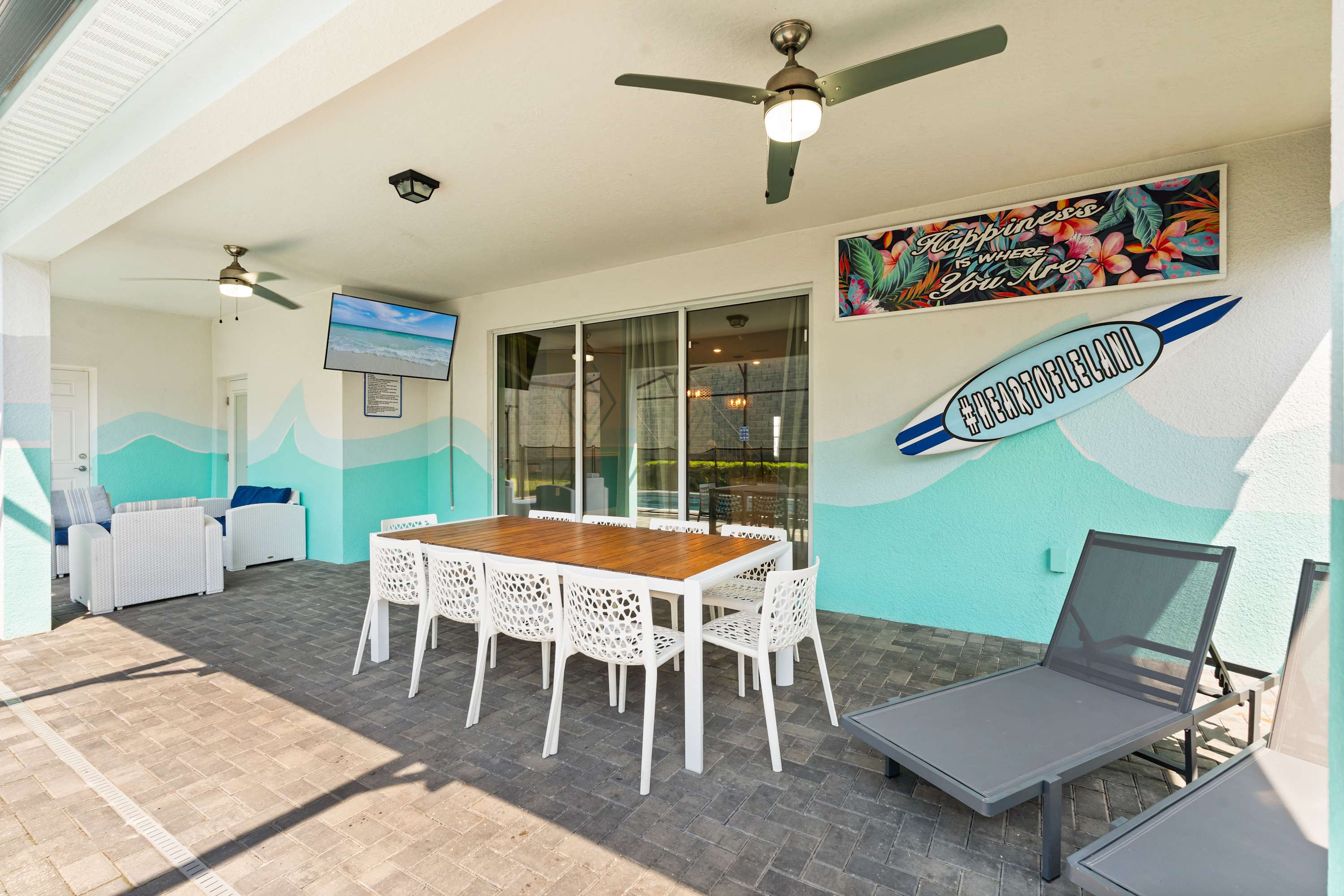 Covered outdoor patio with a large white dining table and chairs, a wall-mounted TV, and beach-themed decor including a surfboard and tropical artwork, overlooking the screened pool area.
