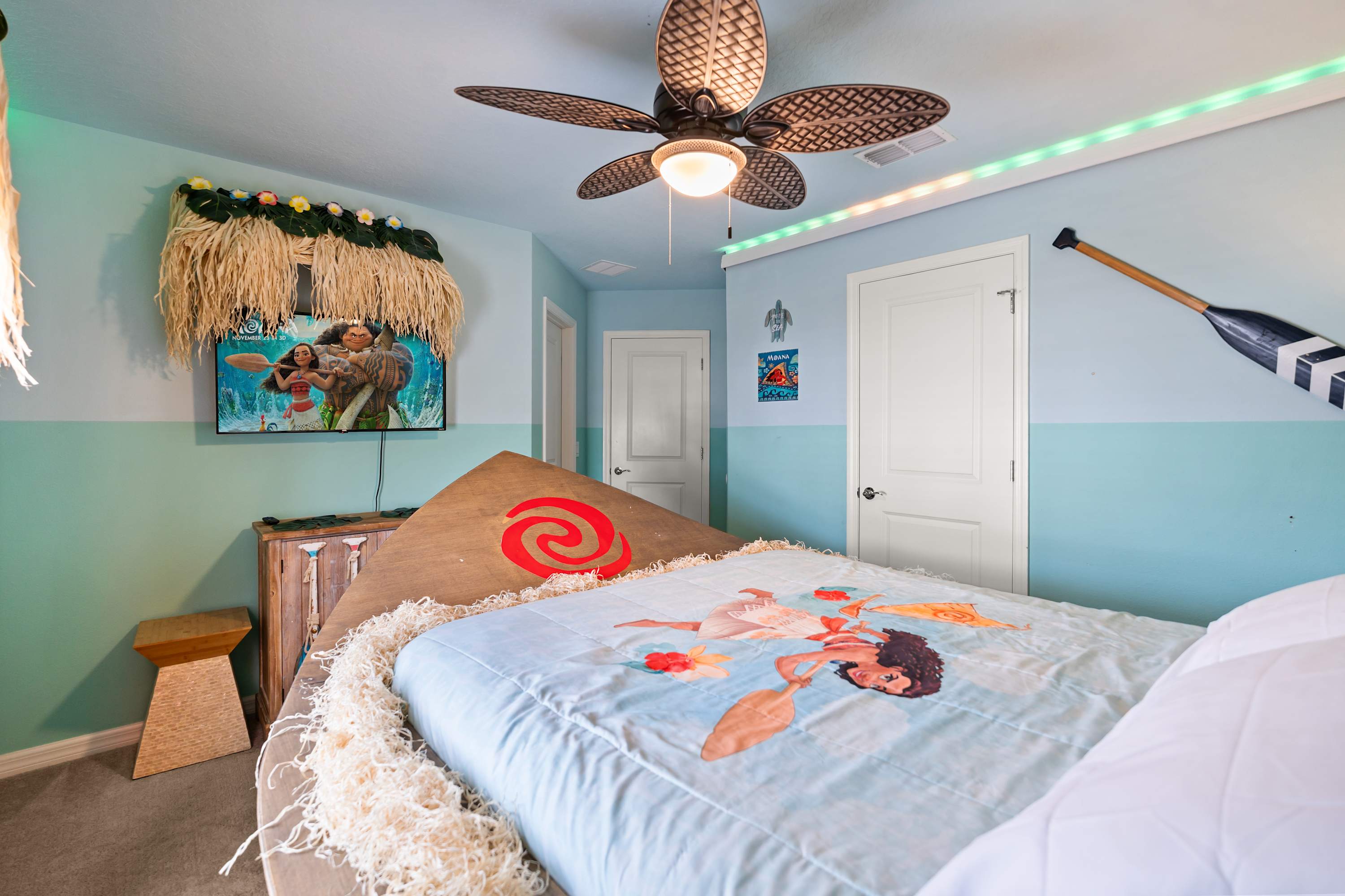 A different angle of the Moana-themed bedroom shows the boat-shaped bed with a Moana-themed comforter, a wall-mounted TV, and a decorative thatched canopy above the TV, with an oar on the wall.