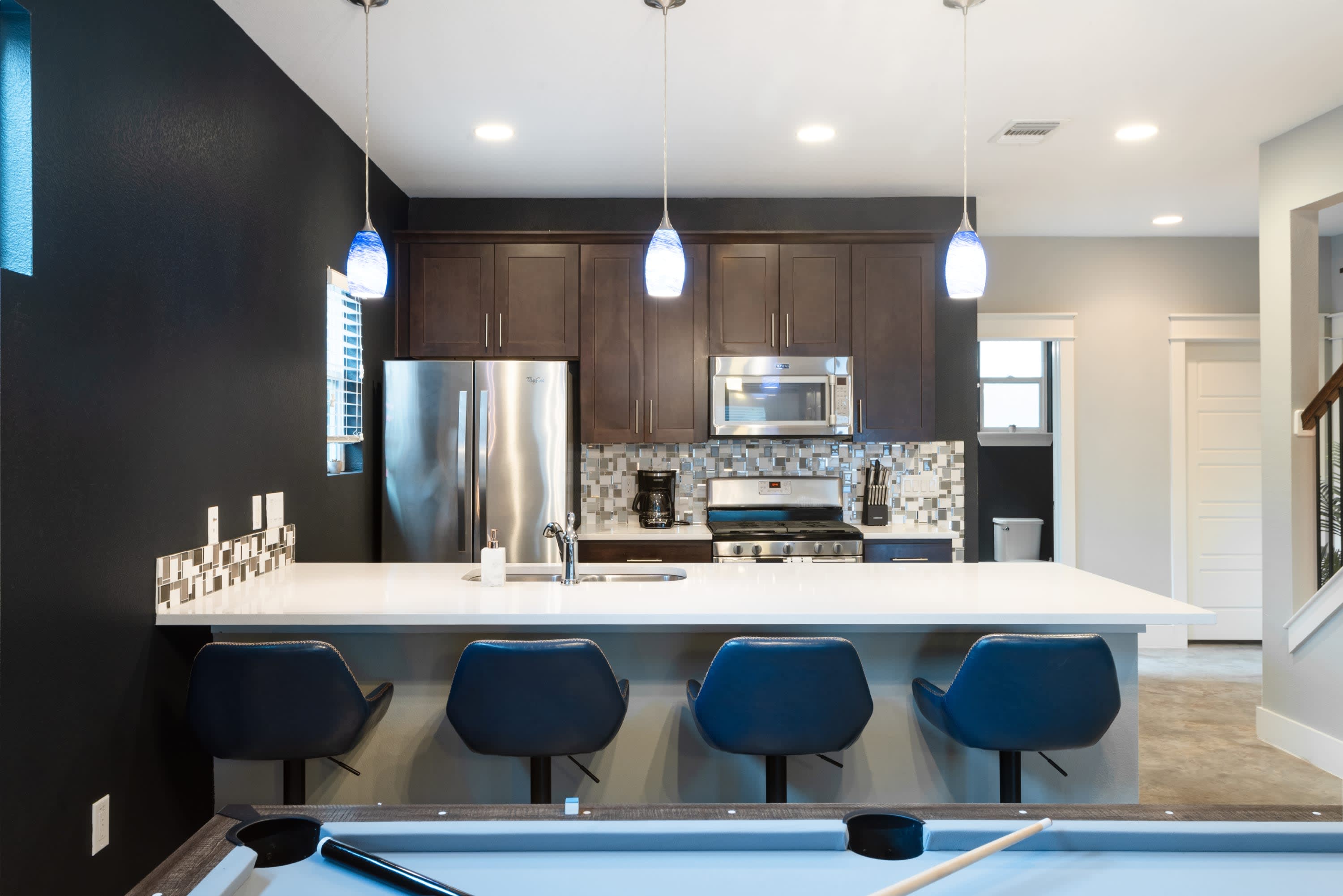 A modern kitchen with dark wood cabinets, stainless steel appliances, and a white quartz island with blue bar stools, viewed from the adjacent game room. Three blue glass pendant lights illuminate the island.