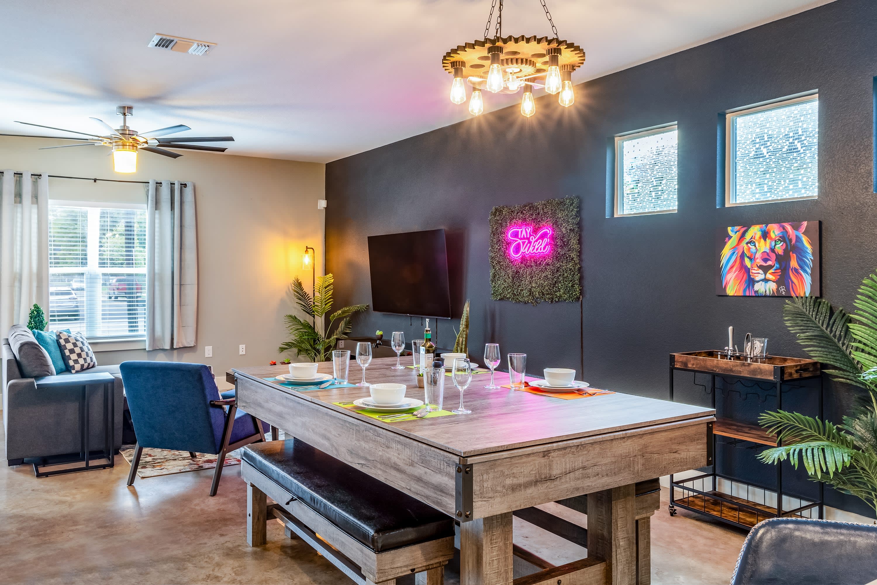 A versatile dining area featuring a large wooden table, which appears to be a convertible pool table, set for a meal with white place settings. The room has a dark accent wall with a "Stay Wild" neon sign and a unique gear-themed chandelier.