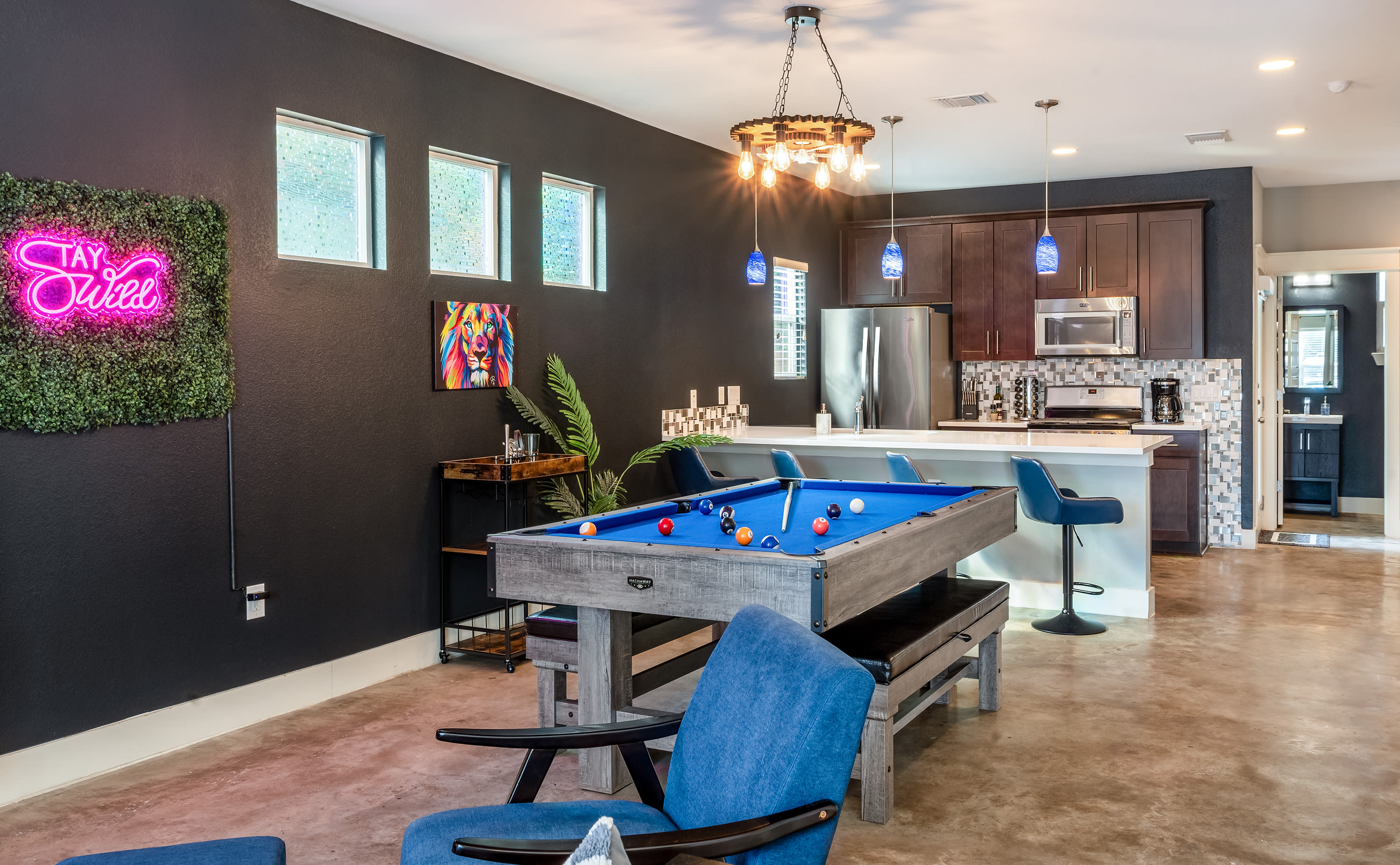 A game room featuring a blue-felt pool table with a wooden frame, set up with billiard balls and cues. The dark accent wall is visible in the background, along with a blue armchair and ottoman.