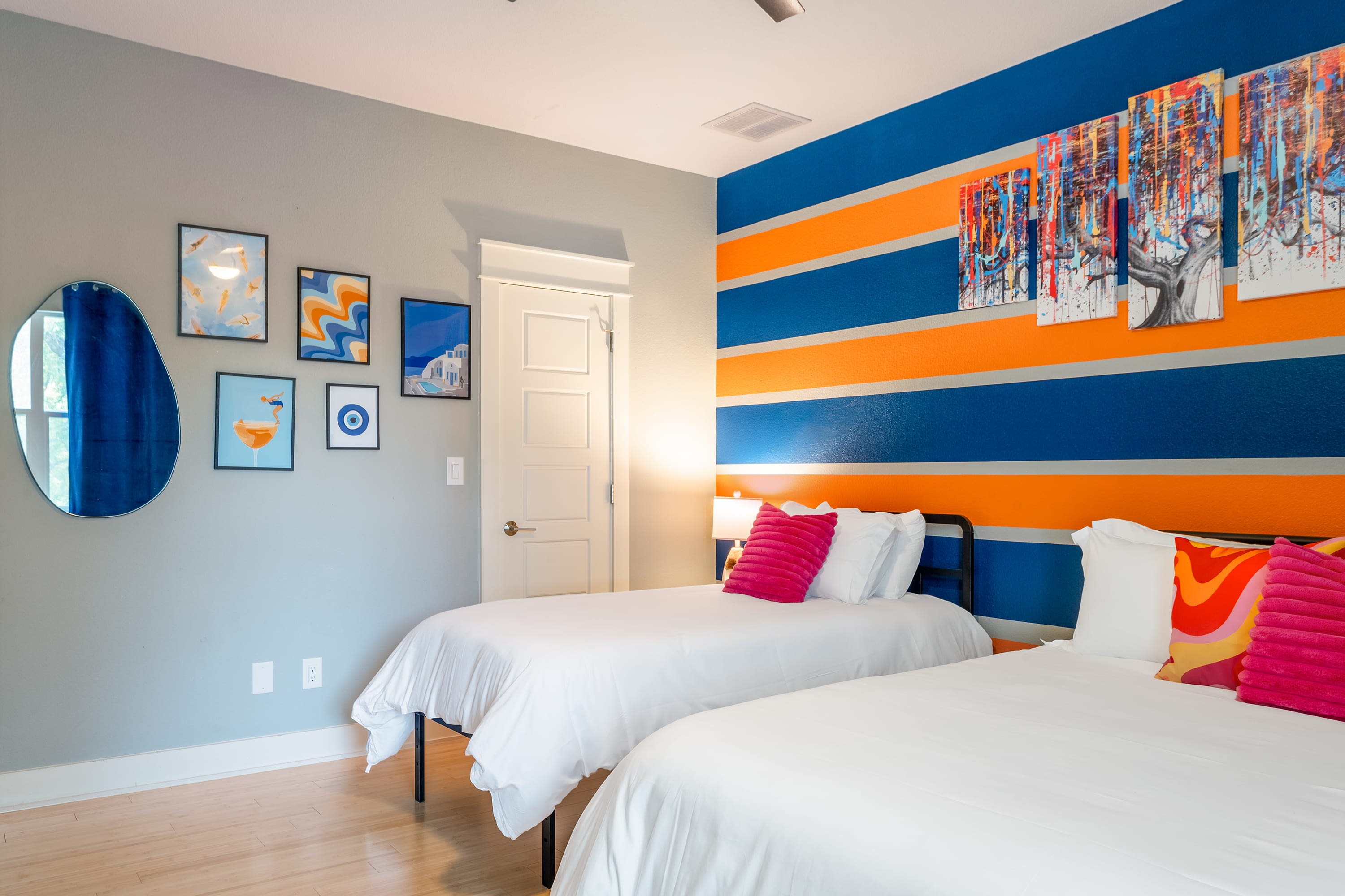 Bright bedroom with light wood floors and a vibrant accent wall featuring bold orange and blue stripes, adorned with colorful abstract art. Two twin beds with white bedding and pink accent pillows are present.