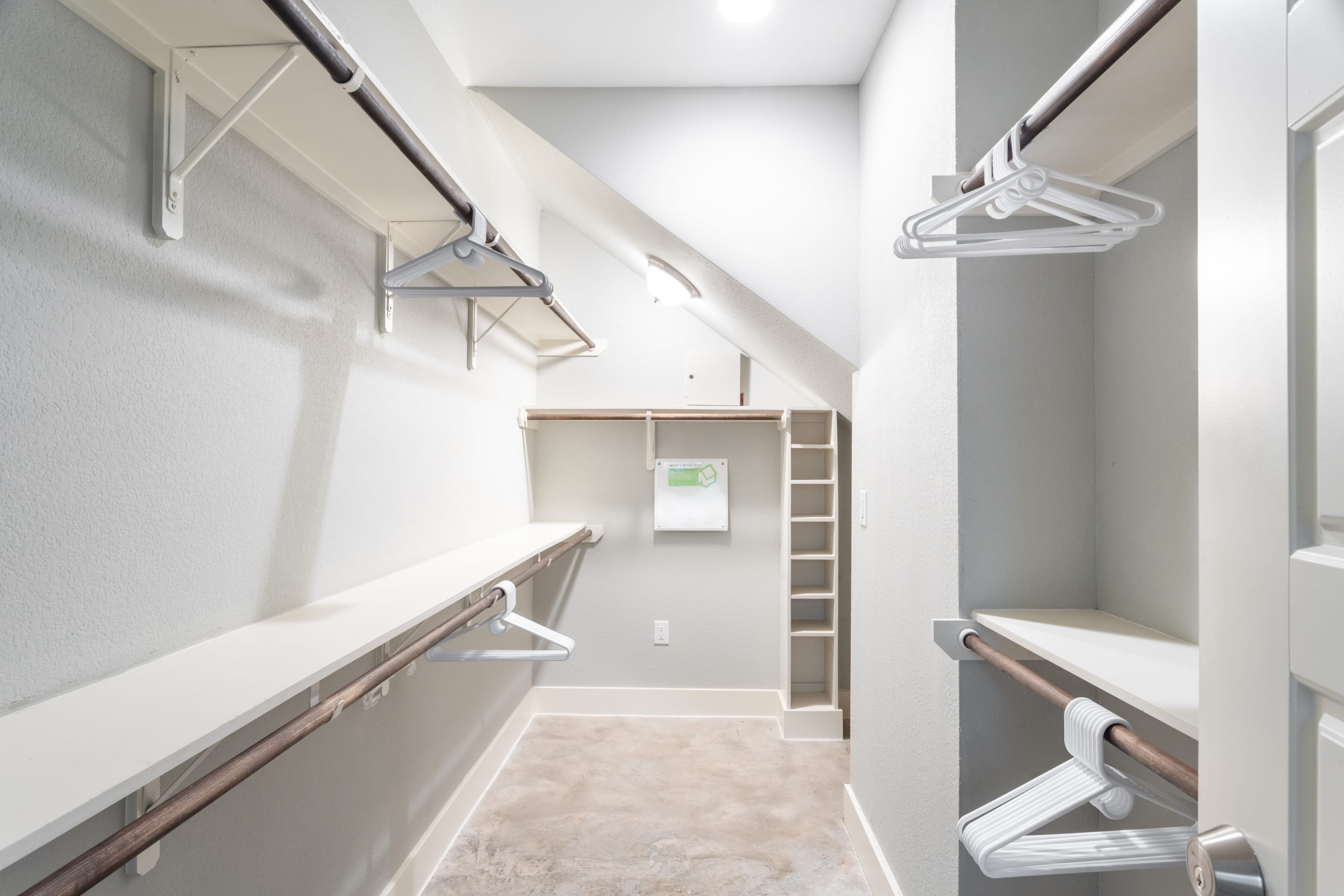 Spacious walk-in closet with white shelving and hanging rods on multiple walls, providing ample storage space. The closet features light gray walls and a concrete floor.