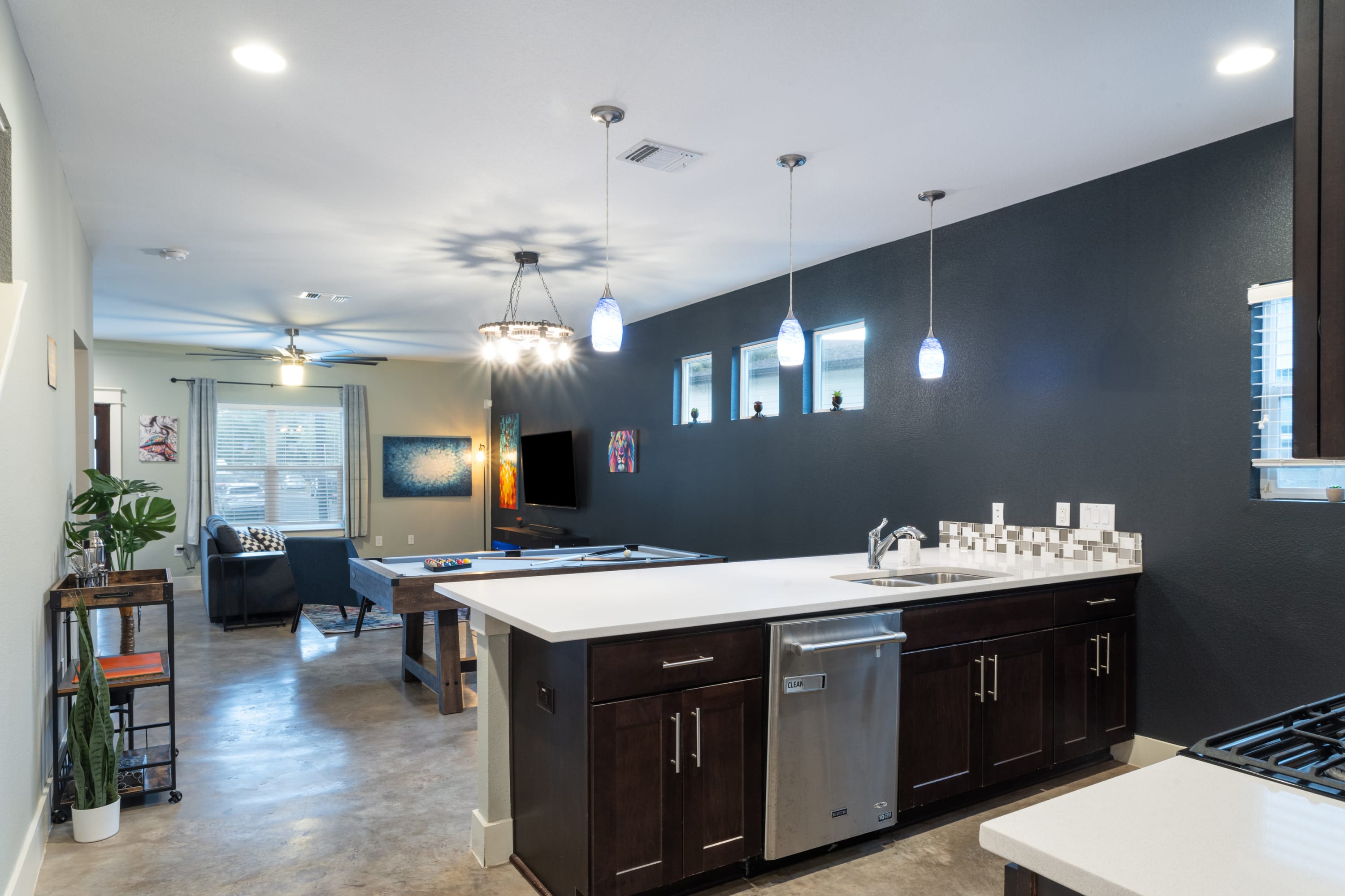 Open-concept entertainment space featuring a modern kitchen island with dark cabinetry and stainless steel appliances, overlooking a living area with a pool table and TV. The room has dark accent walls, concrete floors, and unique pendant lighting.