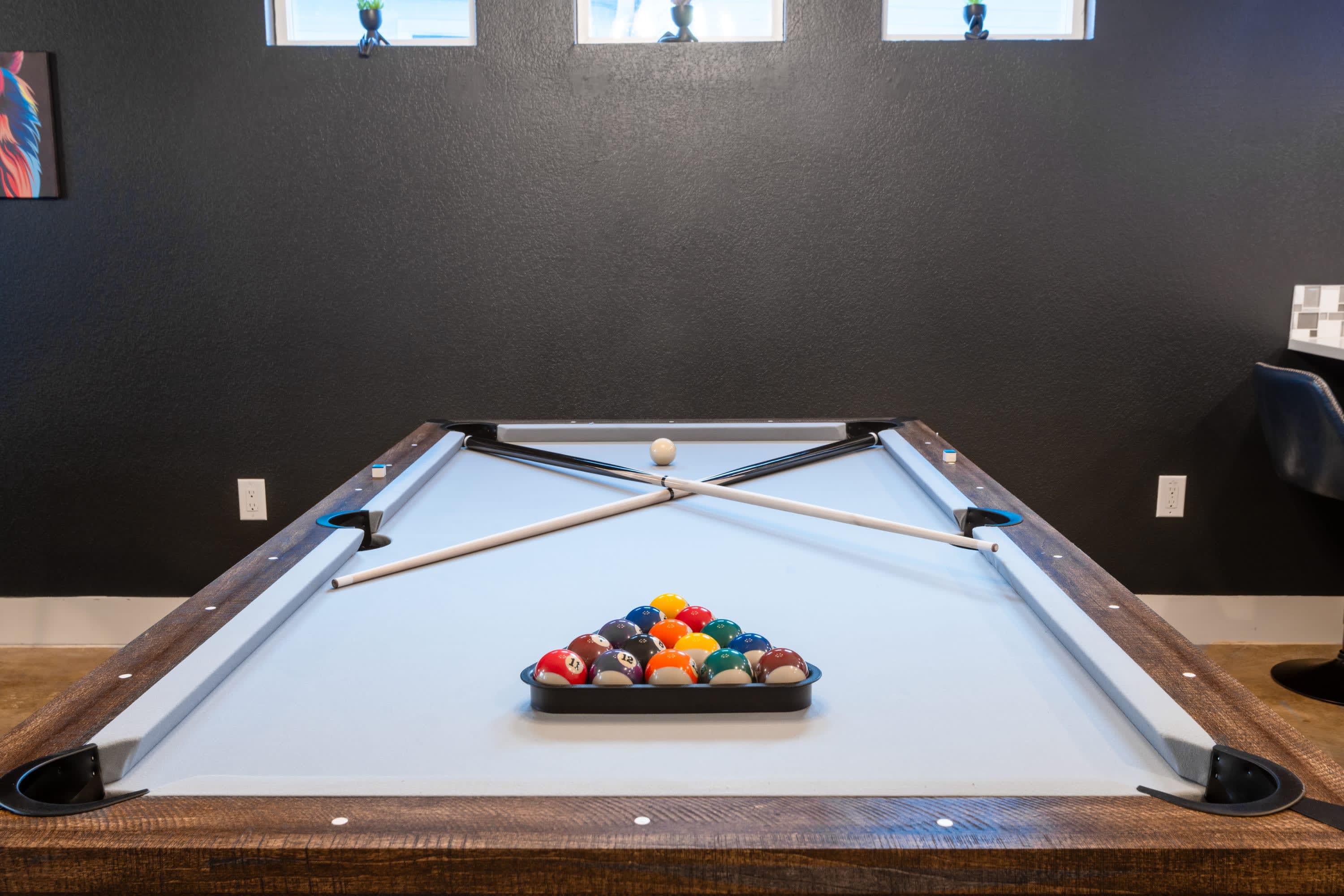 A close-up view of a light blue-felt pool table with a wooden and dark gray frame, featuring a rack of colorful billiard balls and two pool cues crossed on the table.