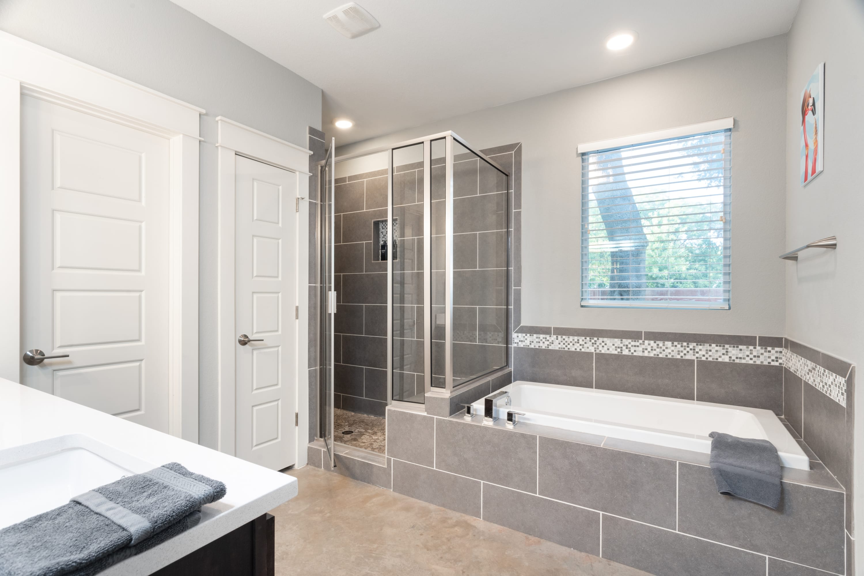 Spacious bathroom with gray tiled floors and walls, featuring a large soaking tub and a separate glass-enclosed walk-in shower. A window with blinds provides natural light, and a double vanity is partially visible.