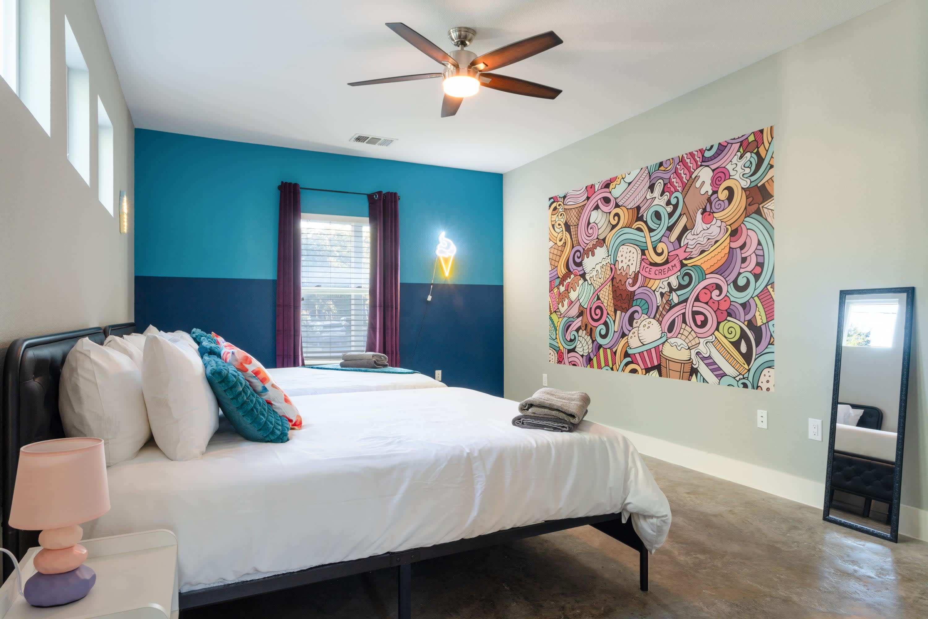 Another view of the vibrant bedroom with concrete floors, showing a large colorful mural on one wall and a full-length mirror. Two queen beds with black frames and teal pillows are visible, along with a bedside table.