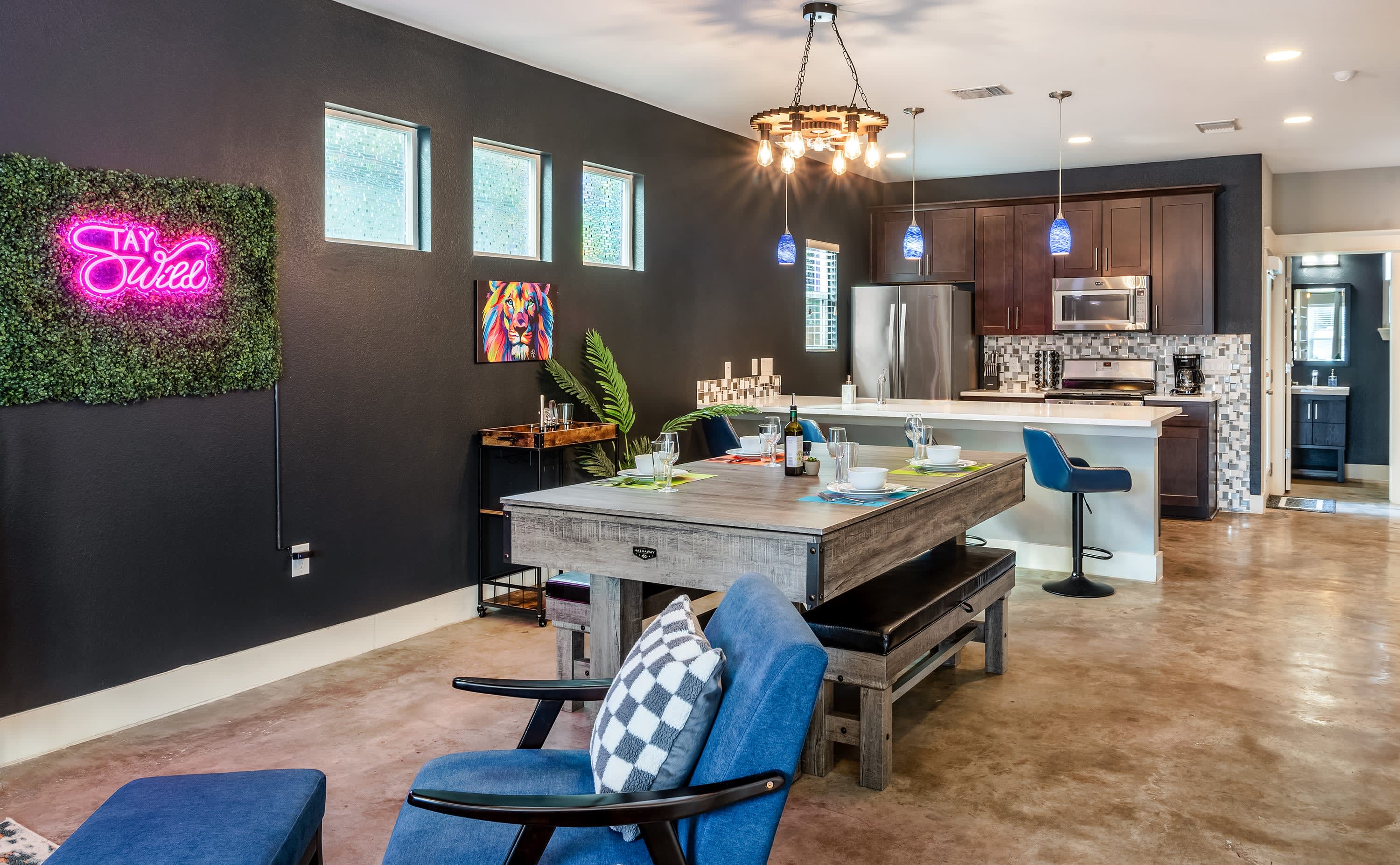 An open-concept living space showcasing a dining table (convertible pool table) in the foreground, leading to a modern kitchen with dark cabinetry and a white island. A dark accent wall features a "Stay Wild" neon sign and a colorful lion art piece.