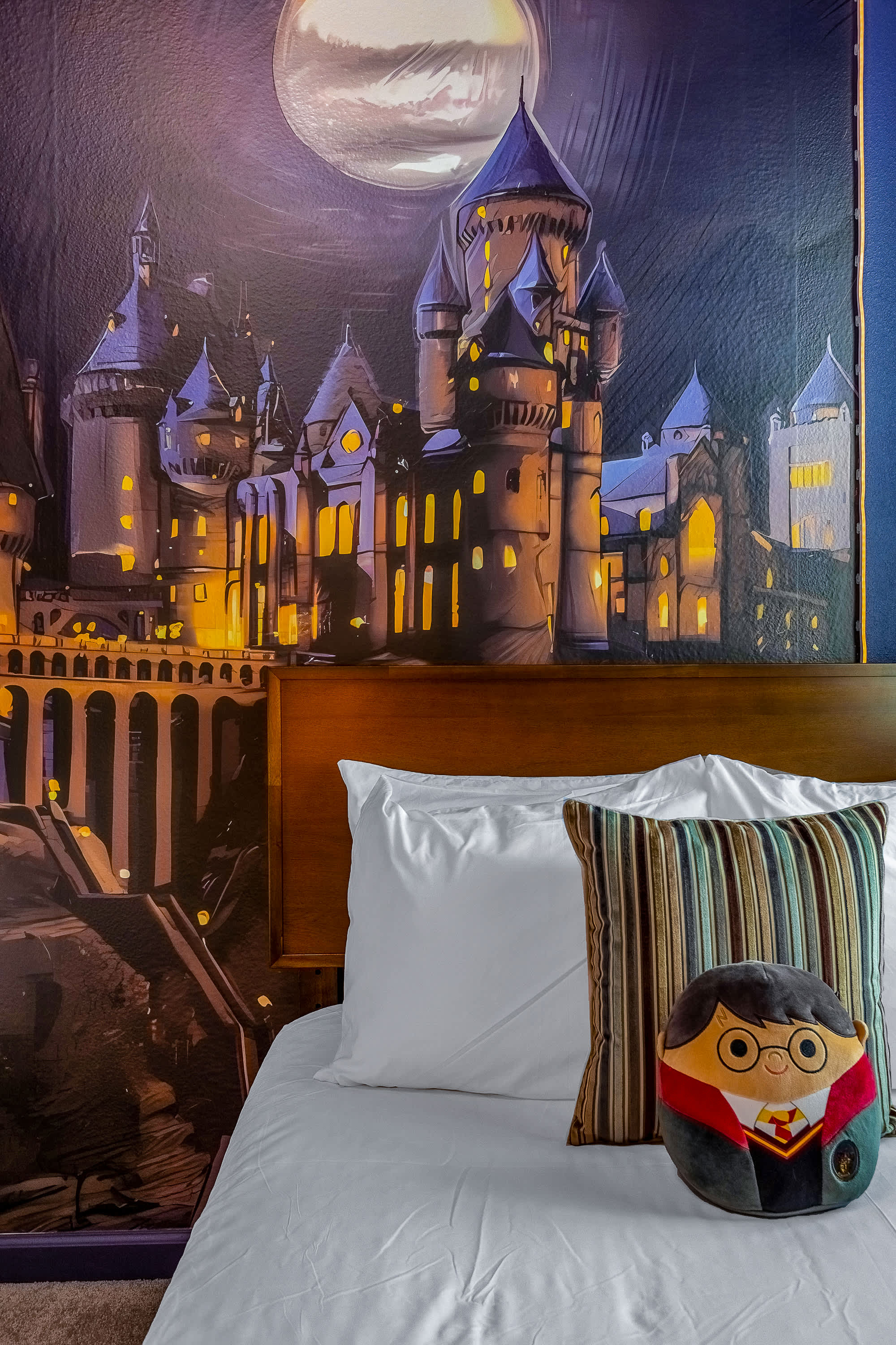 A close-up view of one of the twin beds in the Harry Potter themed bedroom, showing the wooden headboard, white bedding, and a Harry Potter character pillow. The Hogwarts castle mural is prominent in the background.