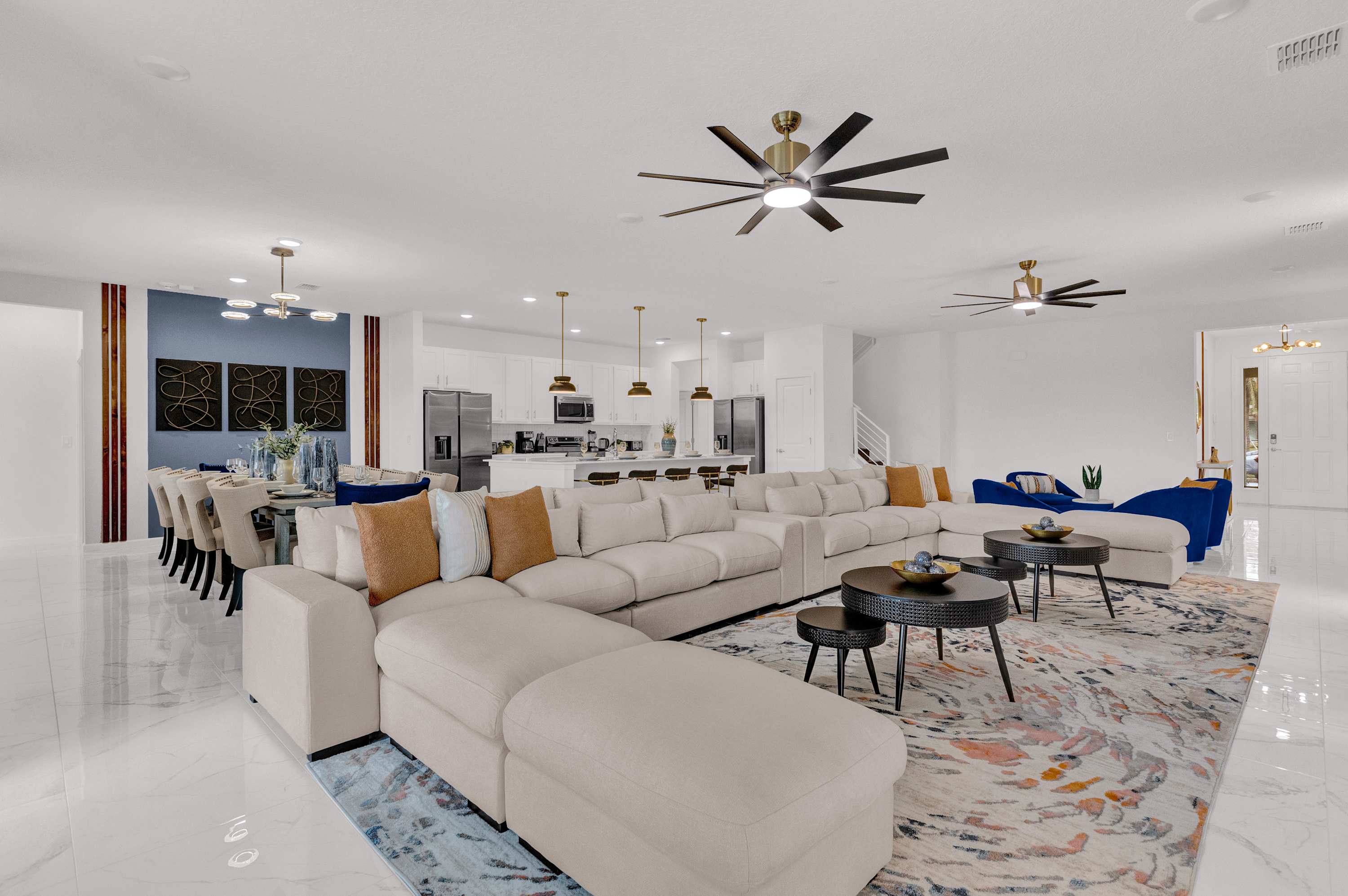 A grand open-concept living room with a long beige sectional sofa, a large area rug, and modern ceiling fans, flowing into a dining area and a contemporary kitchen with white cabinetry.