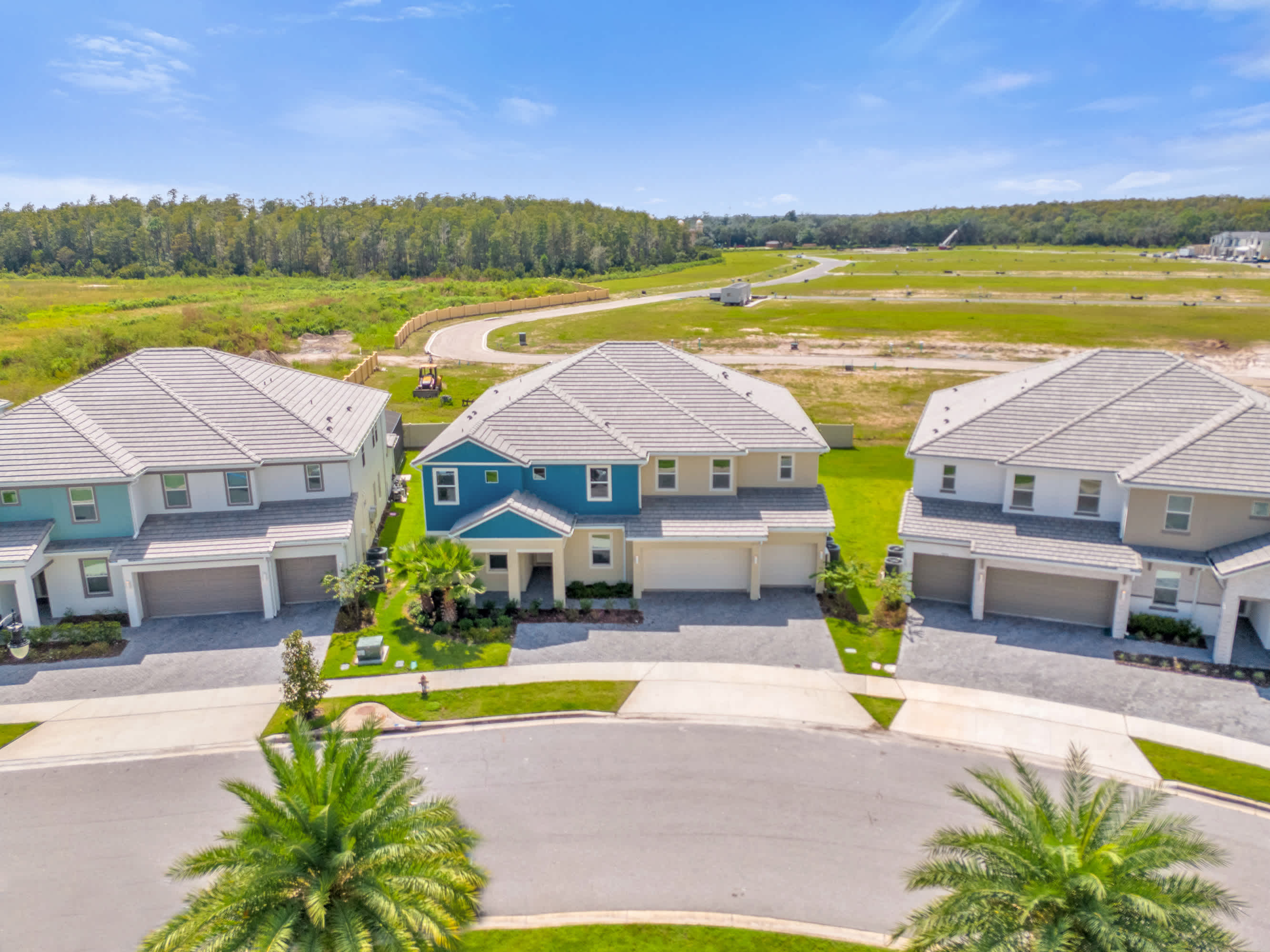 An aerial view showcasing the front of a large, two-story house with a blue and beige exterior, situated in a modern residential community. The house has a two-car garage and is surrounded by paved streets, green lawns, and distant undeveloped land.