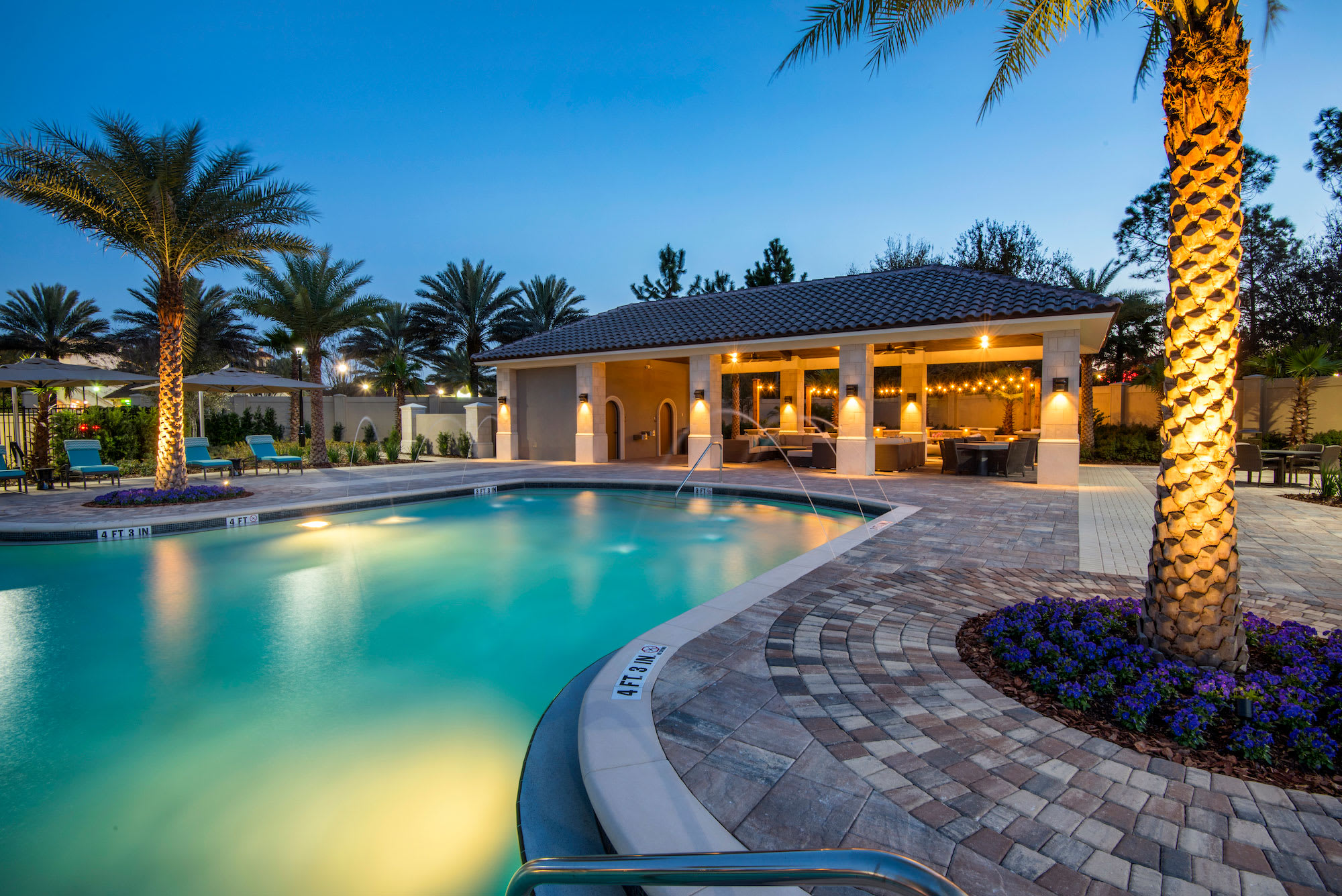 A beautifully lit communal swimming pool at dusk, surrounded by lush landscaping and illuminated palm trees. A covered outdoor lounge area with string lights is visible in the background, creating an inviting evening ambiance.