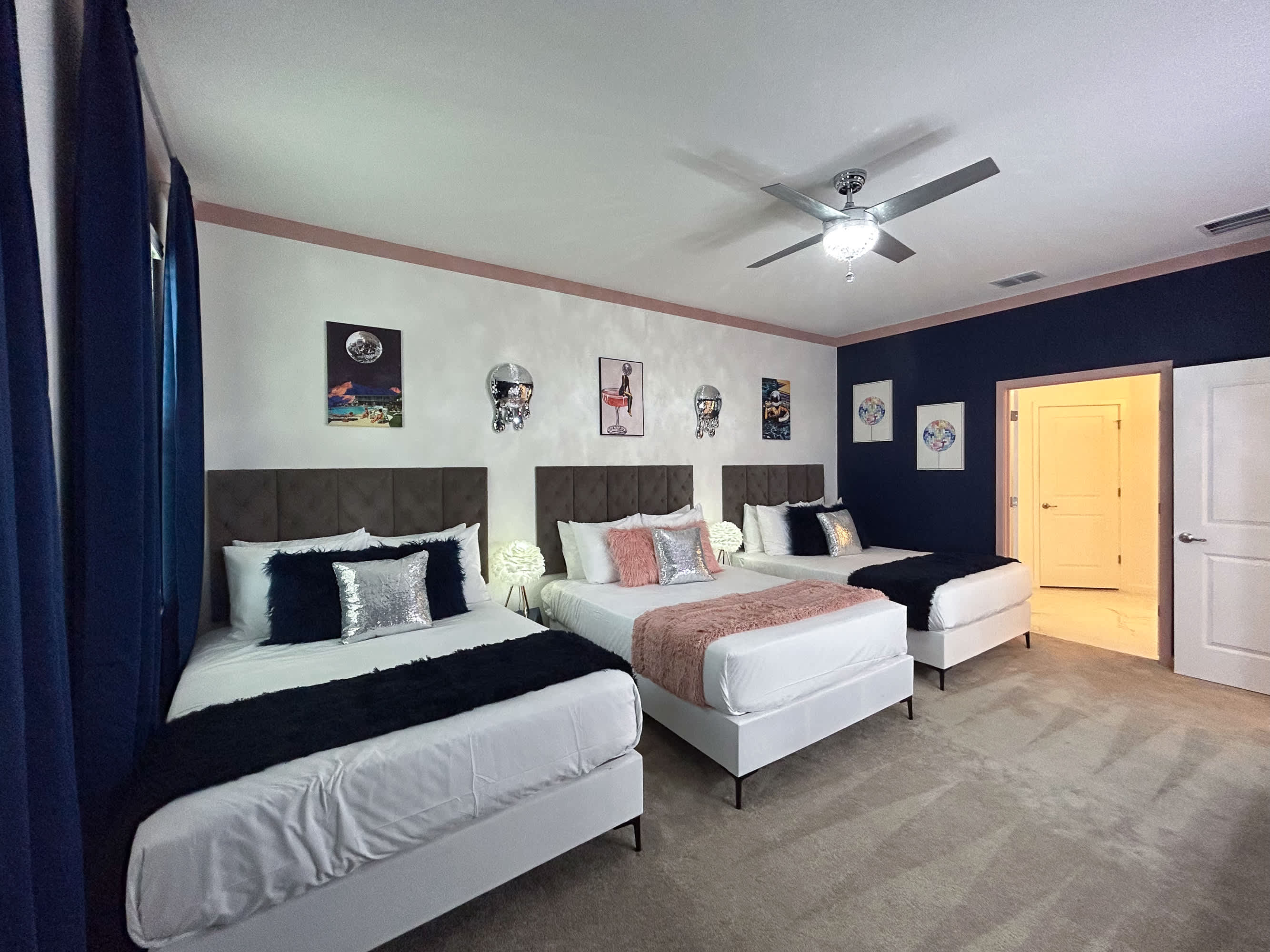 A spacious bedroom featuring three queen-sized beds with upholstered headboards, white bedding, and decorative throws in black and pink. The room has a white and dark blue accent wall, and a ceiling fan.