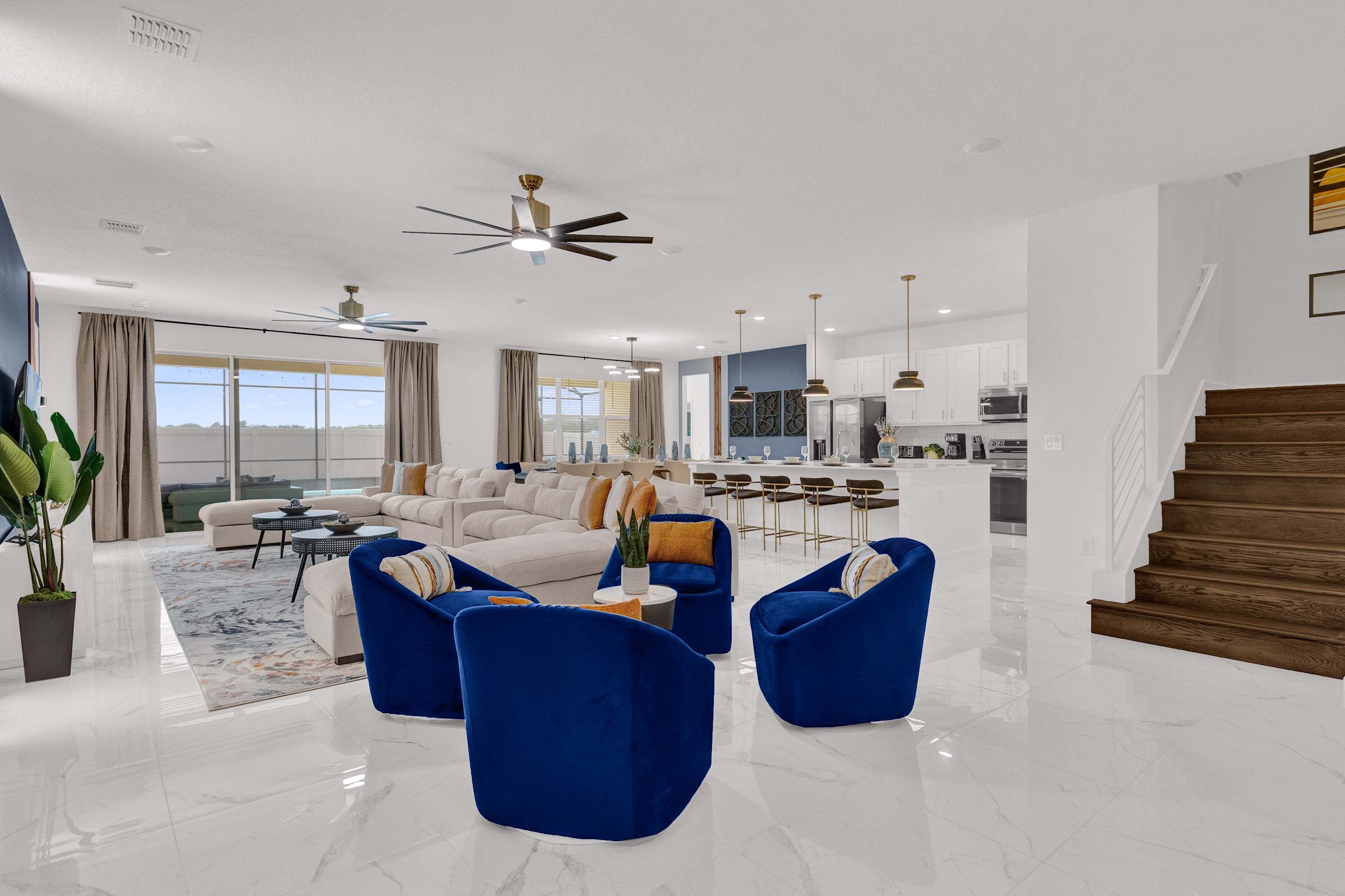 A spacious open-concept living area features white marble-look floors, a large L-shaped sofa, four blue velvet armchairs, and a view towards the kitchen and dining area. Large windows overlook an outdoor space.