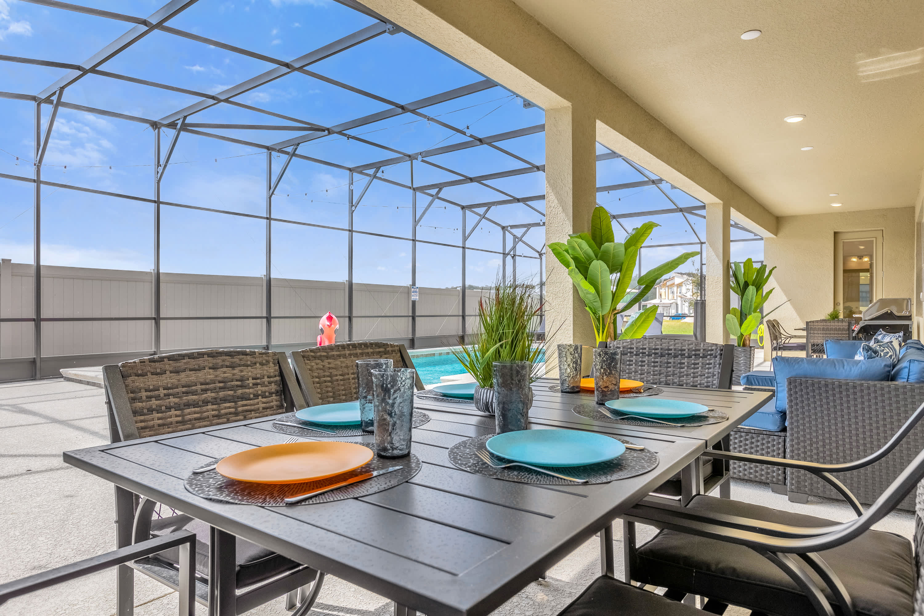 An outdoor dining area on a covered patio, featuring a dark wicker dining table with six chairs, set with blue and orange plates, overlooking a screened swimming pool.