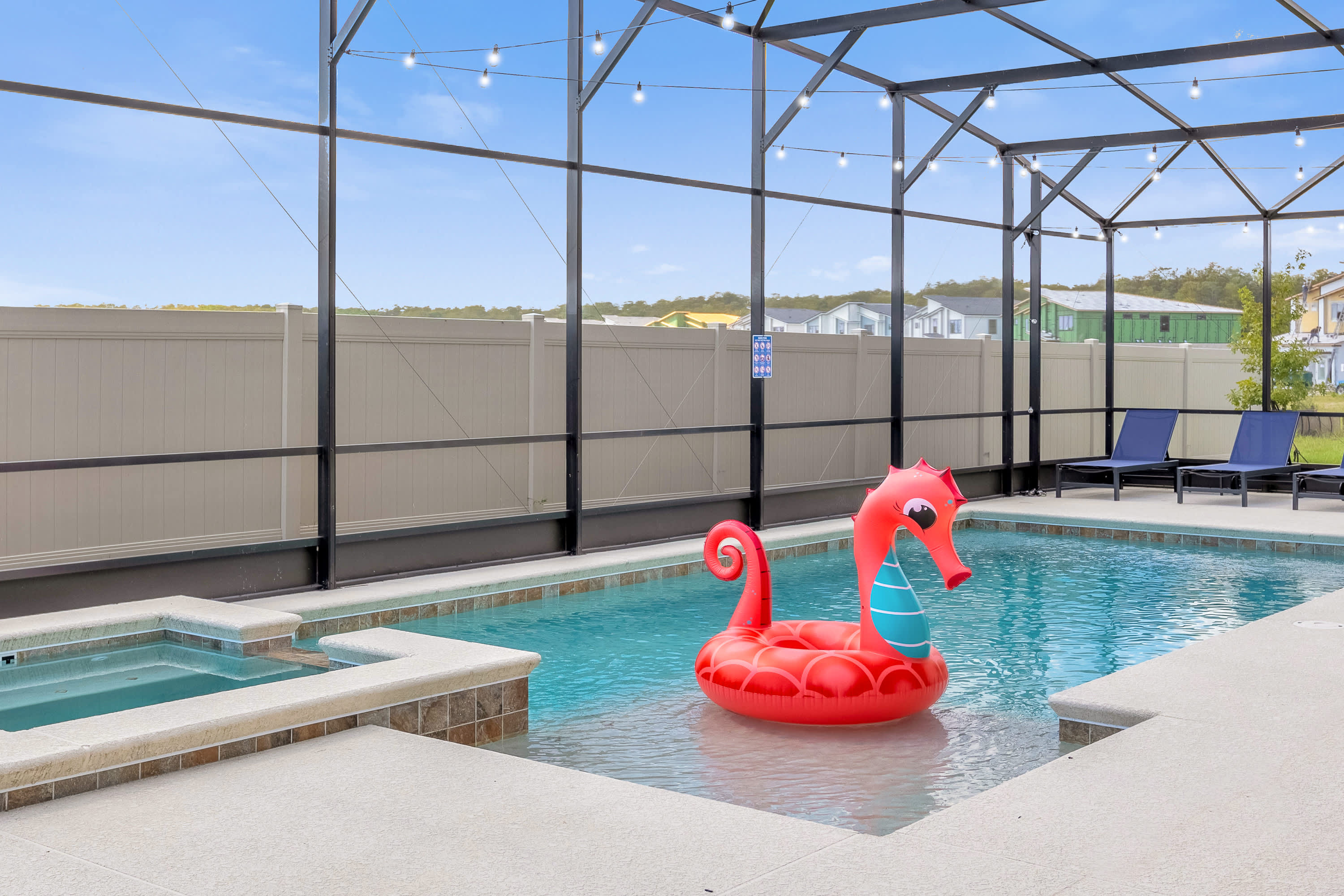 A sparkling blue swimming pool with an integrated hot tub, featuring a large red seahorse float, all enclosed within a screened lanai under a bright sky.