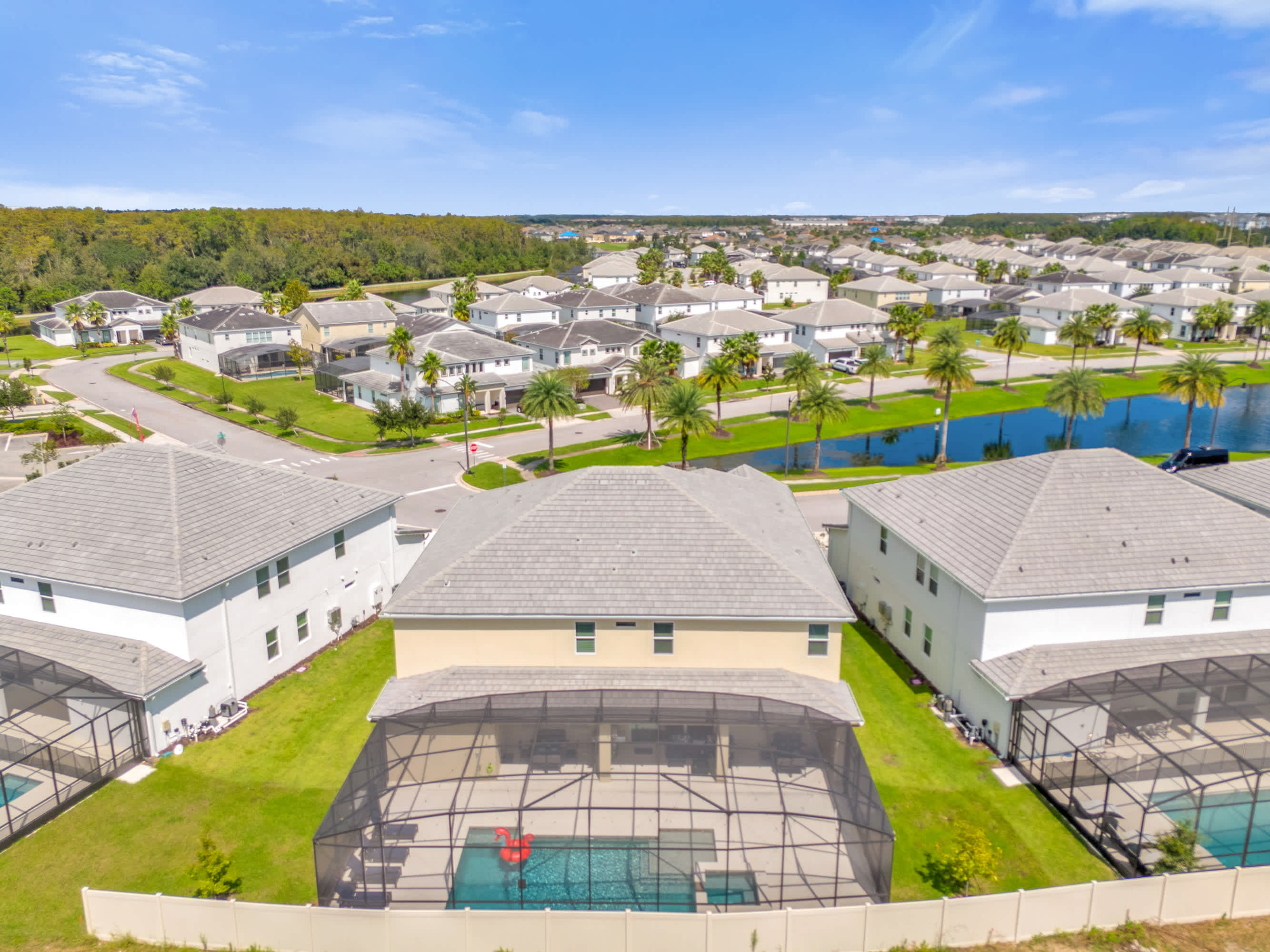 An aerial view of a residential neighborhood with numerous houses, many featuring screened-in private pools, surrounded by green lawns and palm trees. A large pond is visible in the background, under a clear blue sky.
