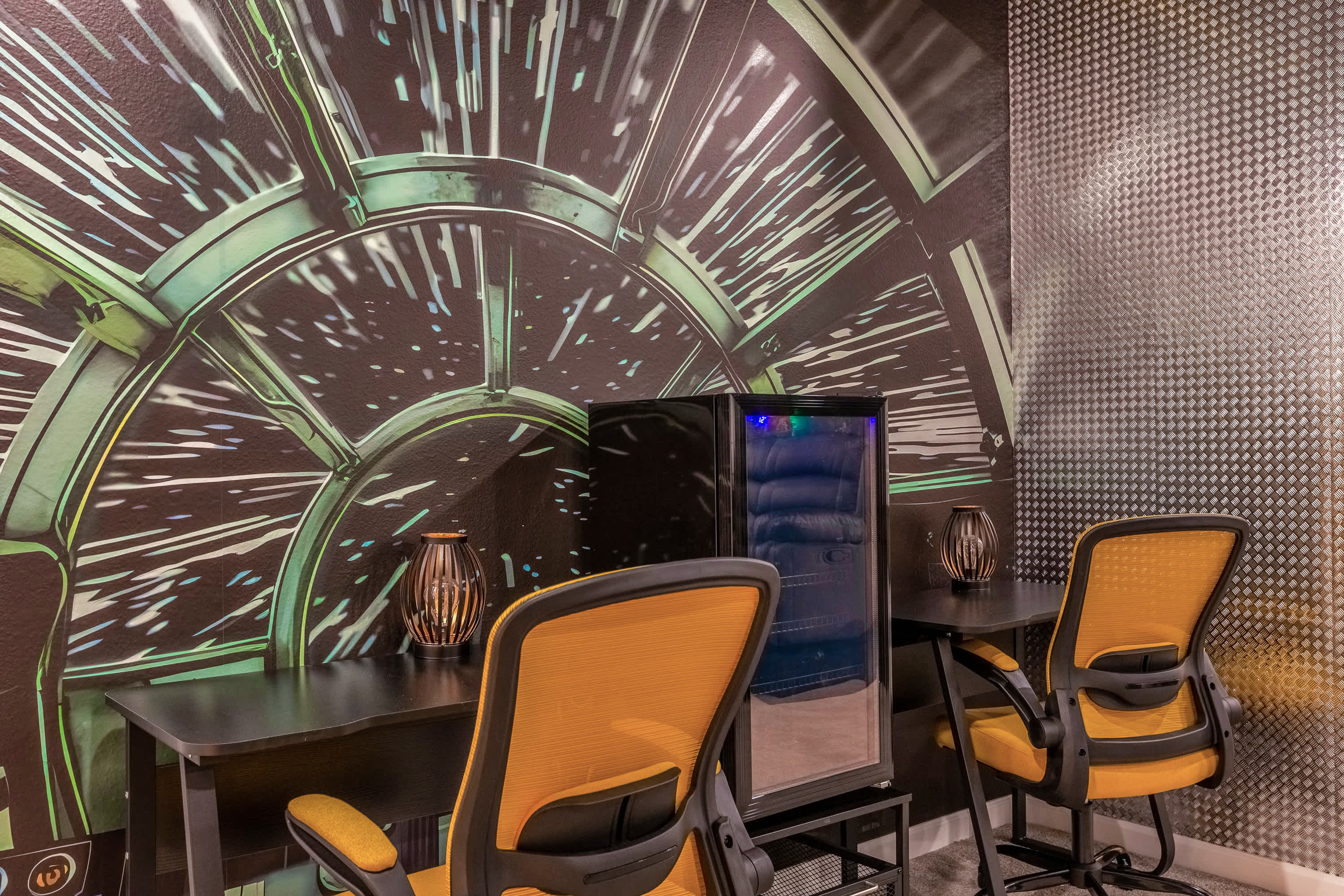 A themed office space with two black desks and orange mesh chairs, featuring a large circular mural on the wall and a mini-fridge, designed for productivity and comfort.