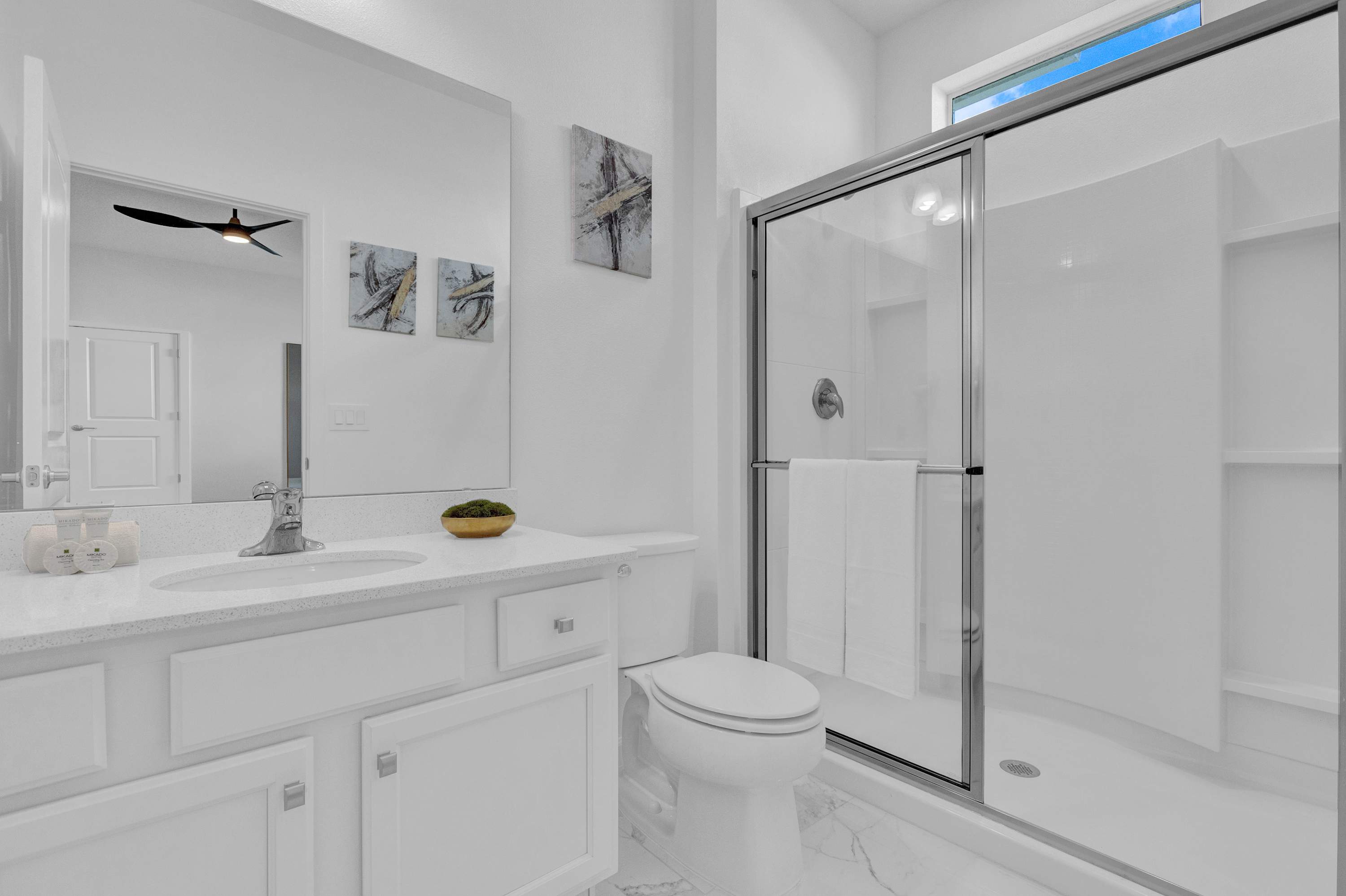 Bright white bathroom featuring a single vanity with a white quartz countertop, a toilet, and a glass-enclosed shower with a clear door. Two abstract art pieces hang above the toilet.