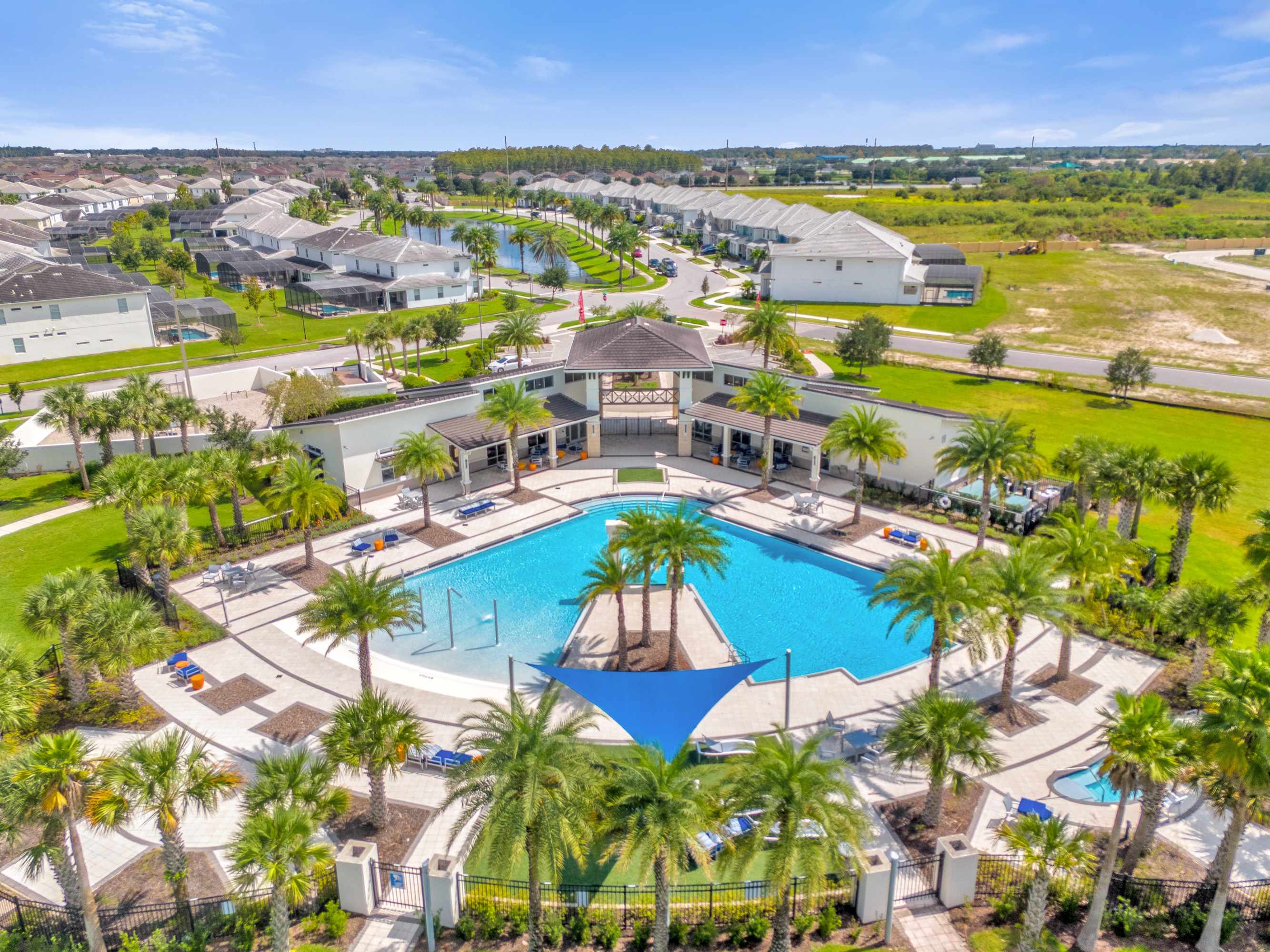 An expansive aerial view of a vibrant community pool area, featuring a large, irregularly shaped swimming pool surrounded by lounge chairs and palm trees. A central clubhouse and shaded seating areas with blue canopies are also visible.