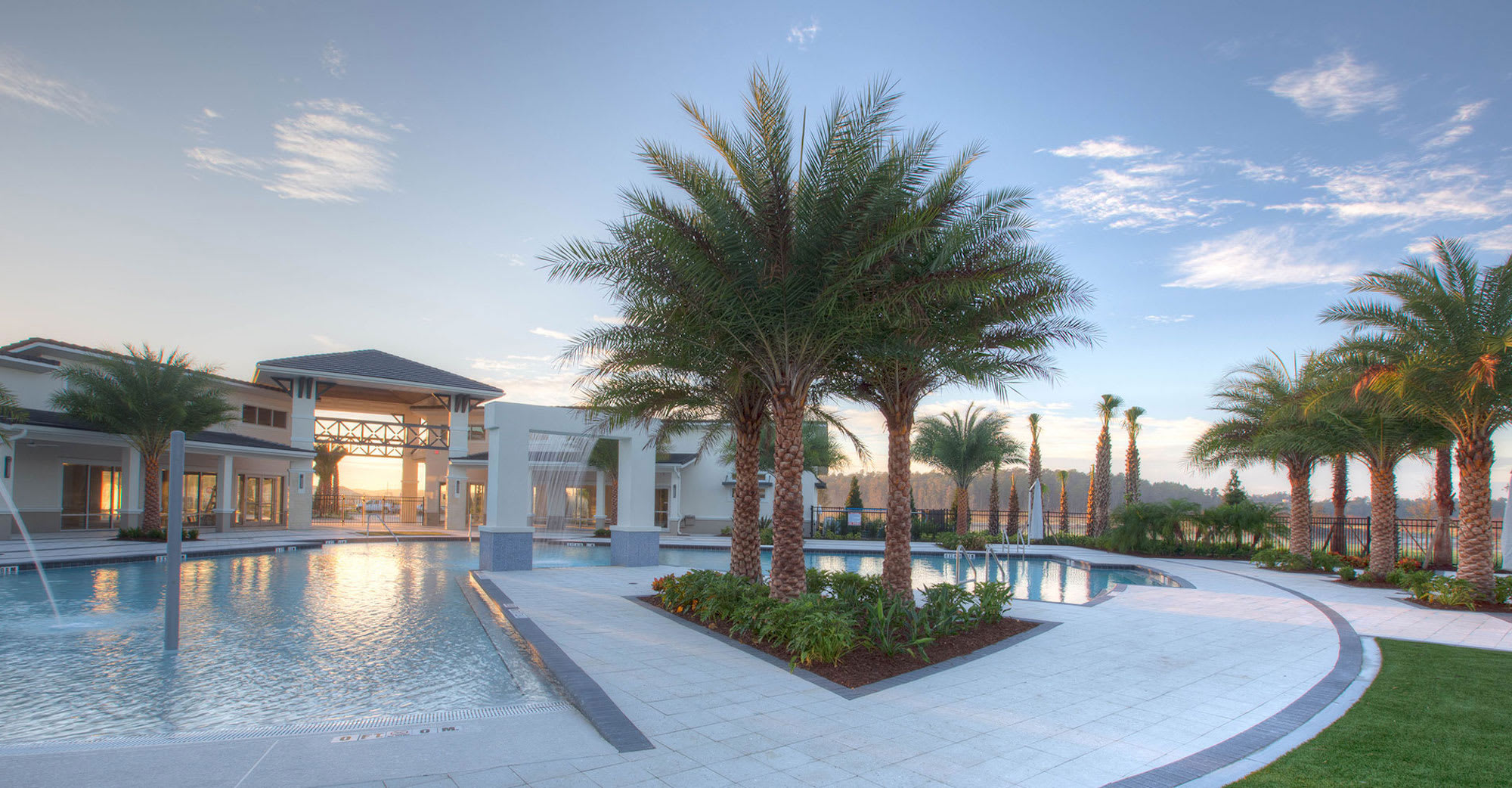 A serene ground-level view of a large communal swimming pool at sunset, featuring multiple palm trees and a modern clubhouse in the background. Water features are active, and the pool deck is illuminated by the warm glow of the setting sun.
