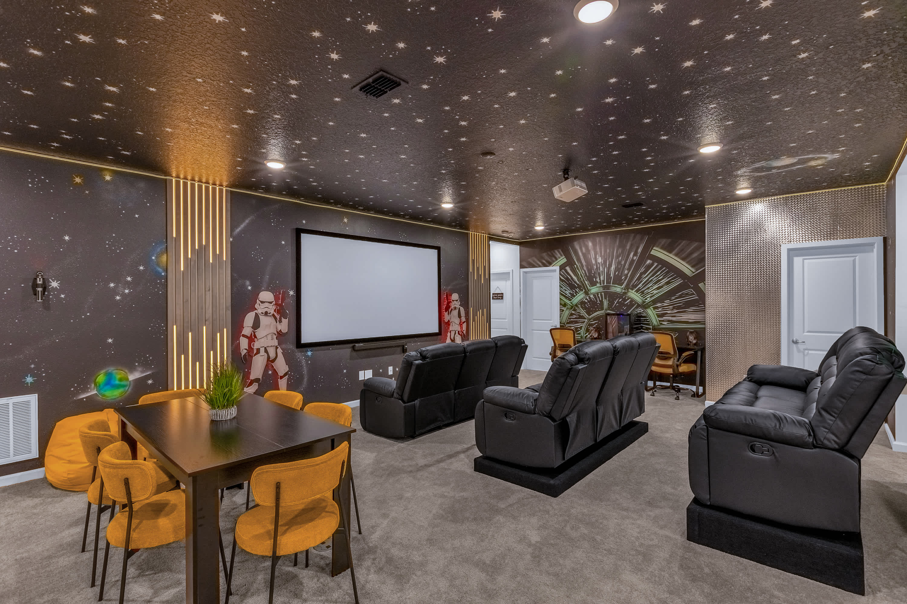 Themed media room with a large projection screen, multiple rows of black reclining sofas, and Star Wars-themed wall murals. The ceiling is dark with star-like lights, and a dining table with yellow chairs is in the foreground.