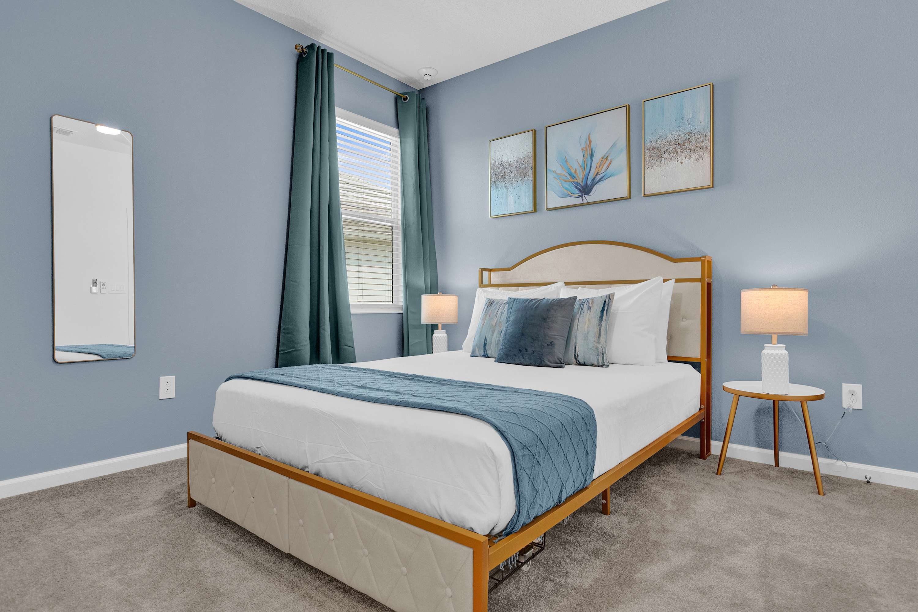 Serene bedroom with light blue walls, a queen bed with a gold and white upholstered frame, and two small round nightstands with white lamps. Three abstract art pieces hang above the bed, and teal curtains frame the window.