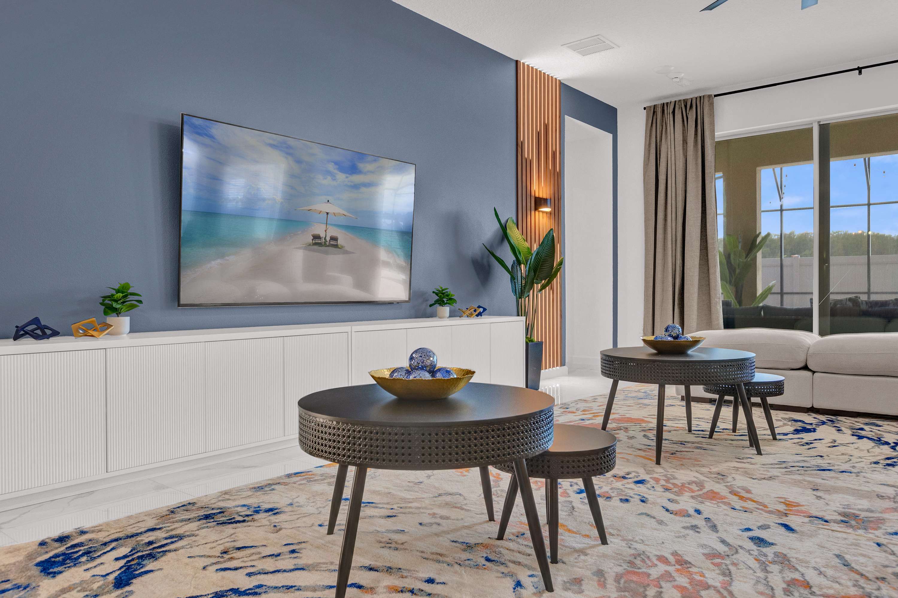 Modern living room with a large wall-mounted TV displaying a beach scene, set against a dark blue accent wall. A long white media console sits below the TV, flanked by decorative wooden slats and a plant.