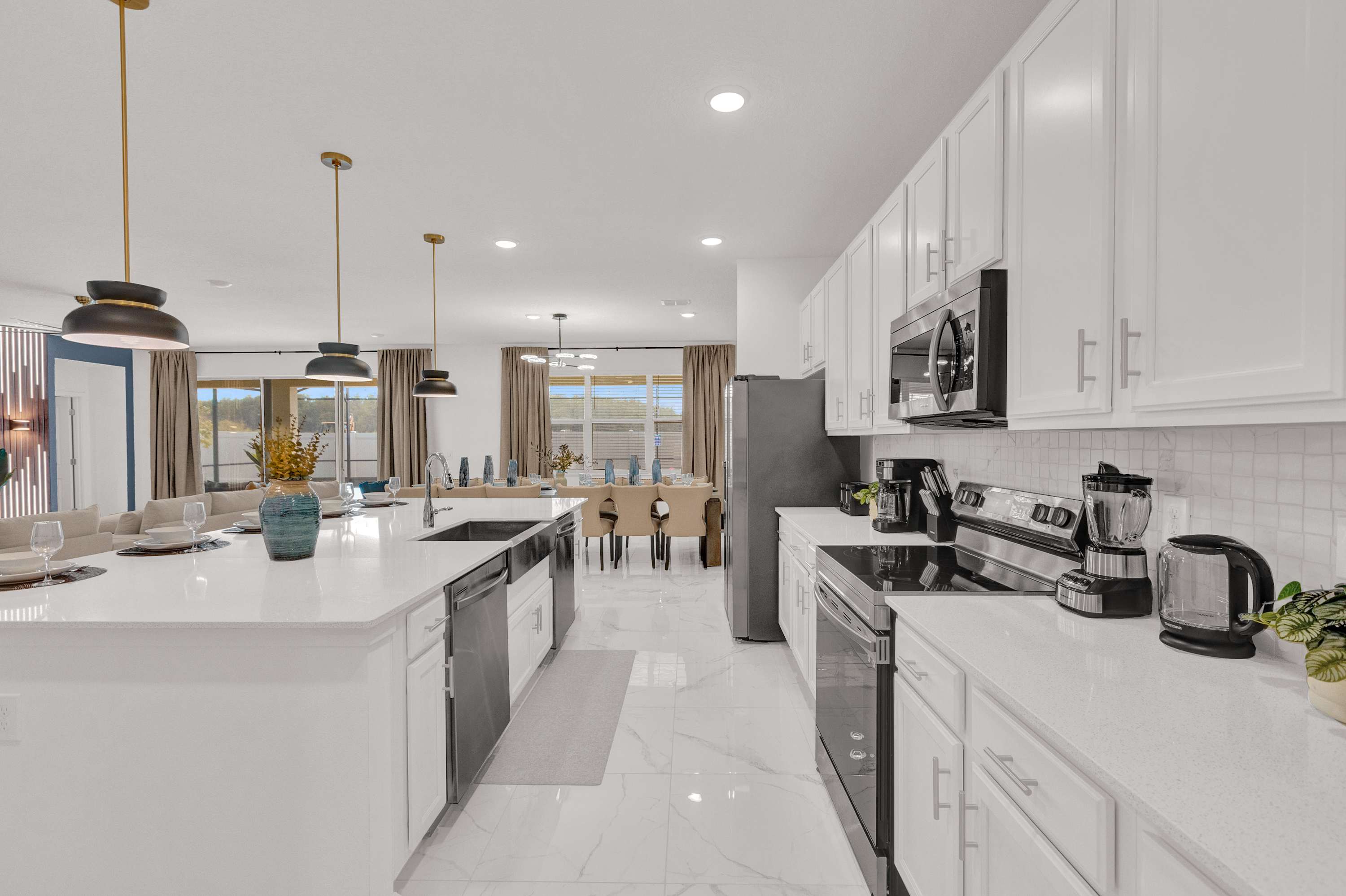 Contemporary kitchen featuring white cabinetry, stainless steel appliances including a stove and microwave, and a large island with a sink. The space has white marble-look floors and opens to the dining and living areas.