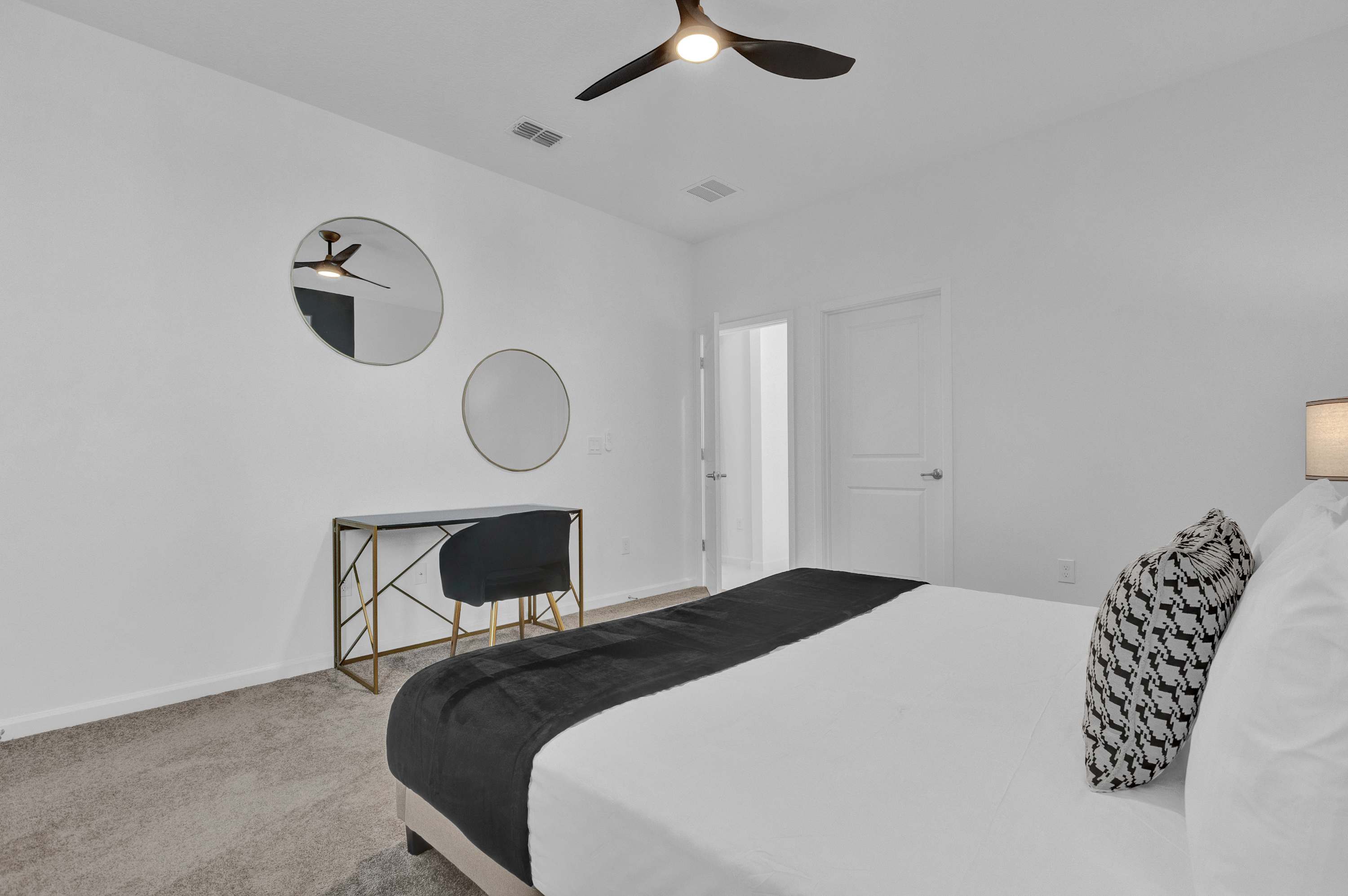 View of bedroom-4 showcasing a black and gold desk with a black velvet chair, two round decorative mirrors on the white wall, and a ceiling fan, offering a chic and functional space.