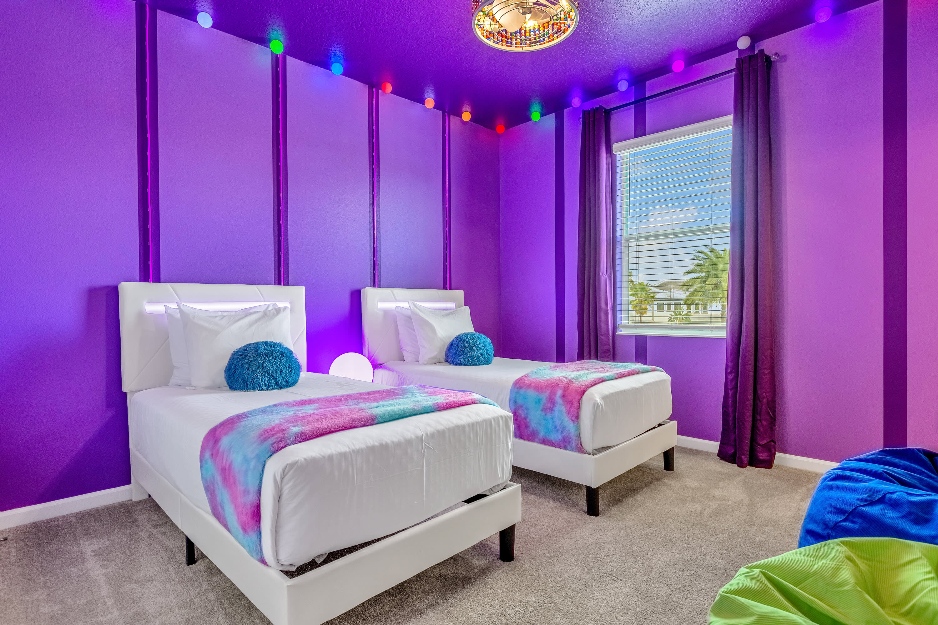 A vibrant purple-themed bedroom with two white-framed beds, colorful string lights adorning the walls, and a large window with white blinds, creating a playful and bright atmosphere.