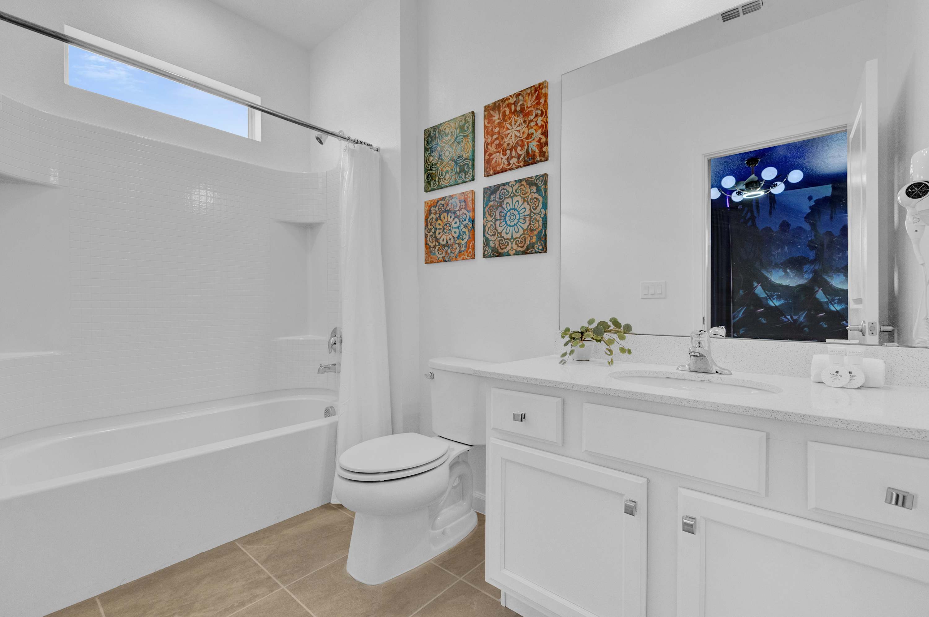 A bright white bathroom with a single vanity, a large mirror, and a toilet. A white tiled tub and shower combo is on the right, with a horizontal window above it. Colorful square art pieces adorn the wall.