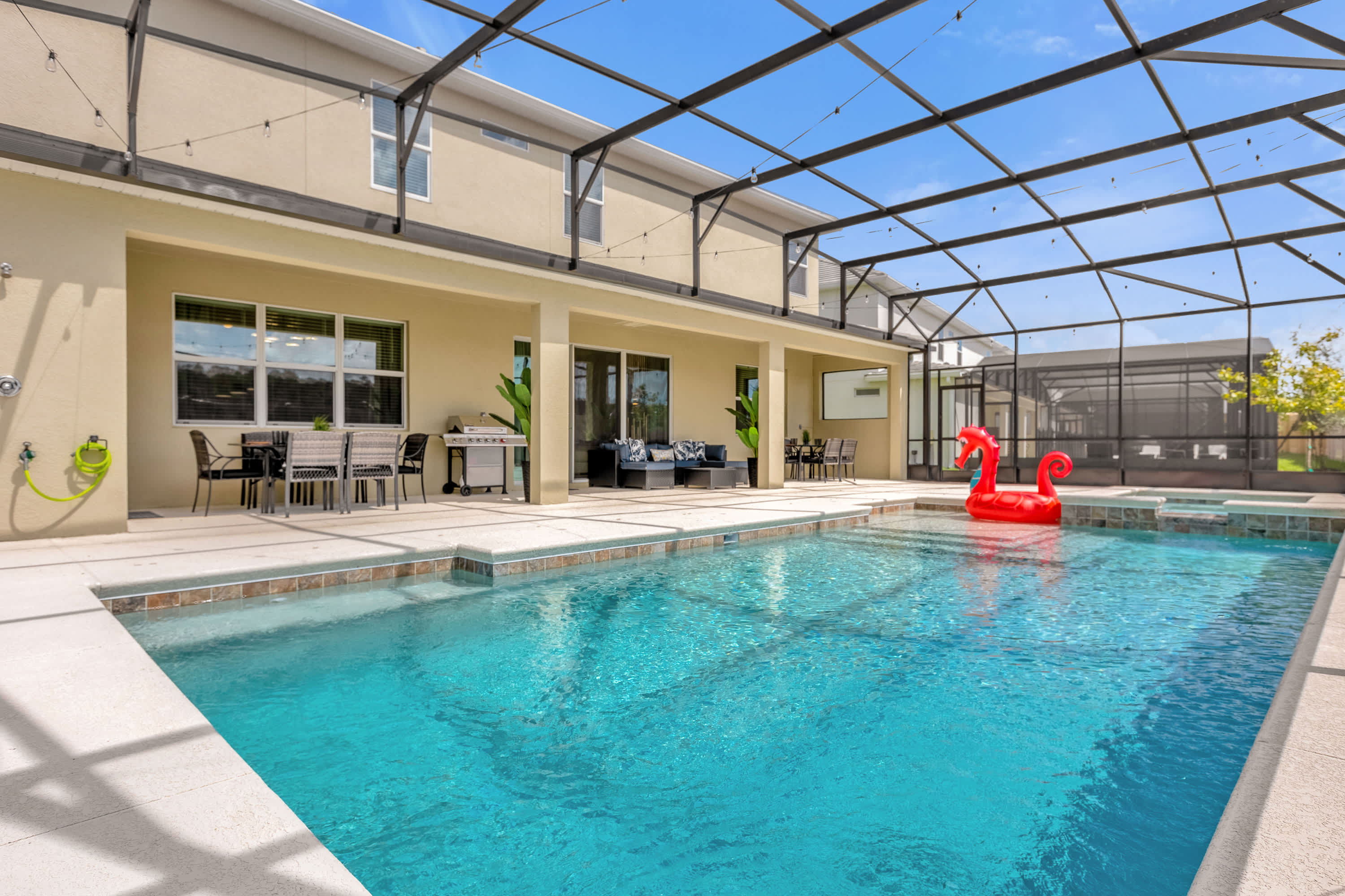A large screened outdoor pool area with a rectangular swimming pool, an integrated hot tub, and a spacious patio with outdoor dining and lounge furniture, viewed from across the pool.