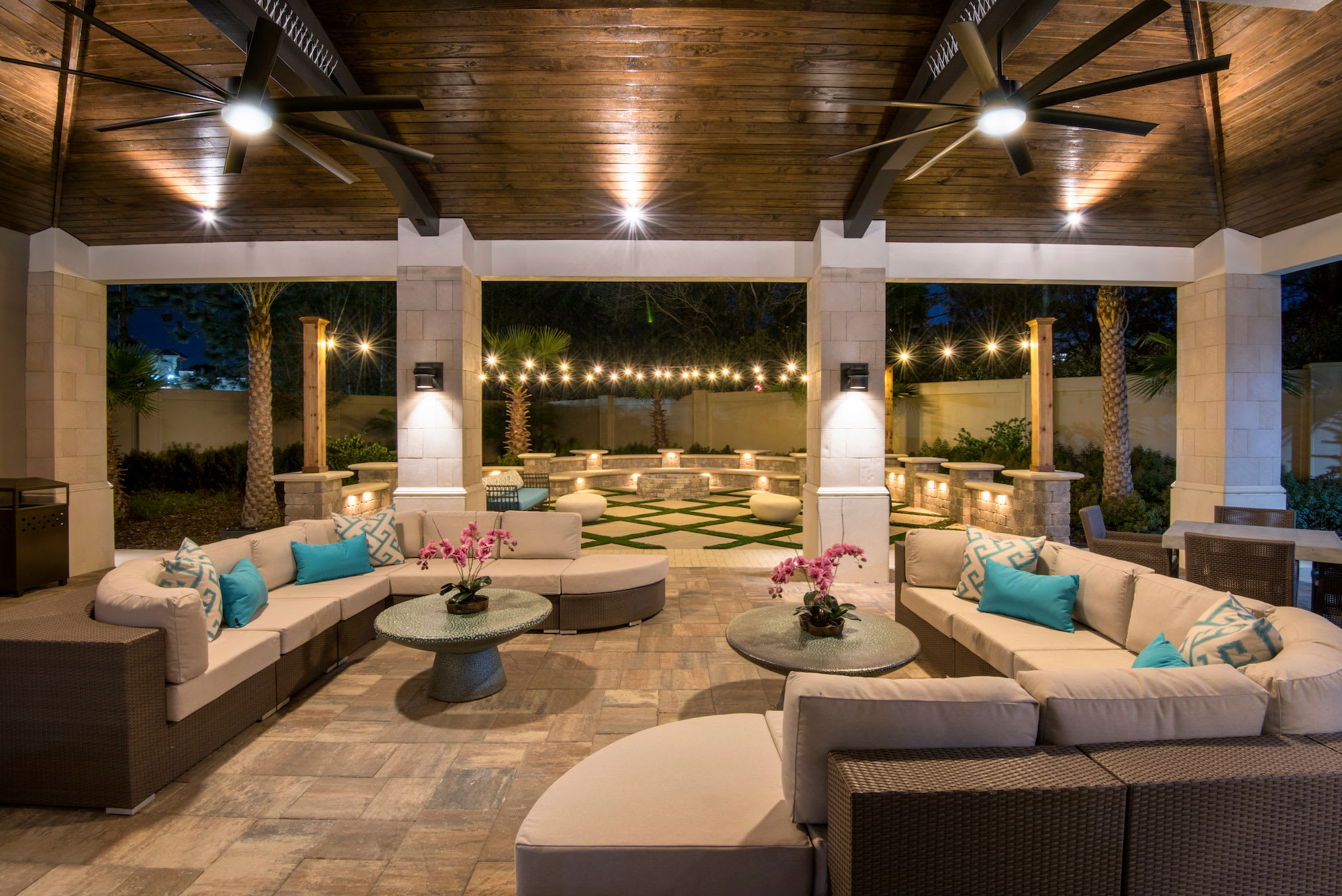 The covered outdoor patio is illuminated at night, showcasing comfortable sectional sofas with teal pillows and round coffee tables on a patterned stone floor. String lights create a festive ambiance, leading to a fire pit area in the landscaped background.