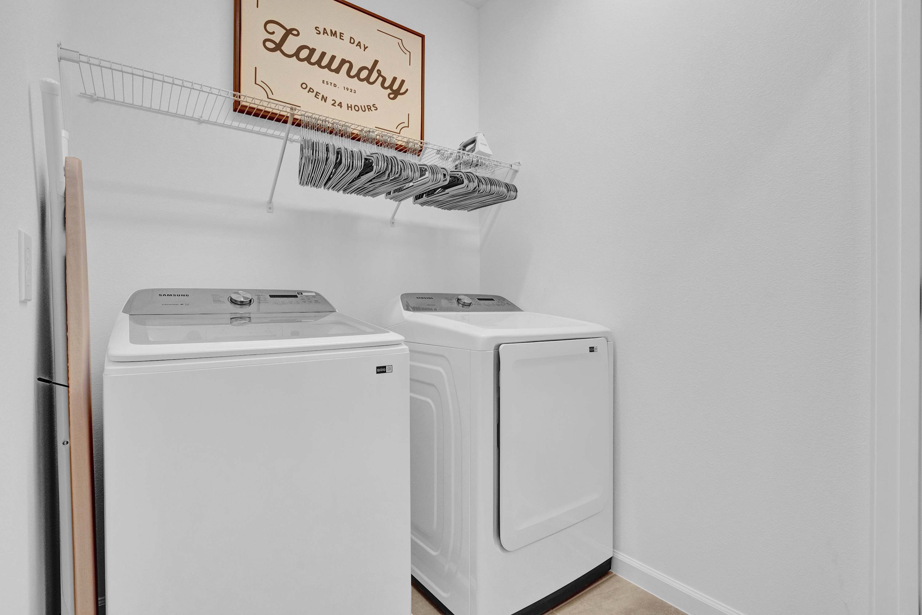 A bright laundry room with white walls, featuring a top-loading washing machine and a front-loading dryer, both in white. A wire shelf above holds numerous hangers, and a decorative 'Same Day Laundry' sign hangs on the wall.