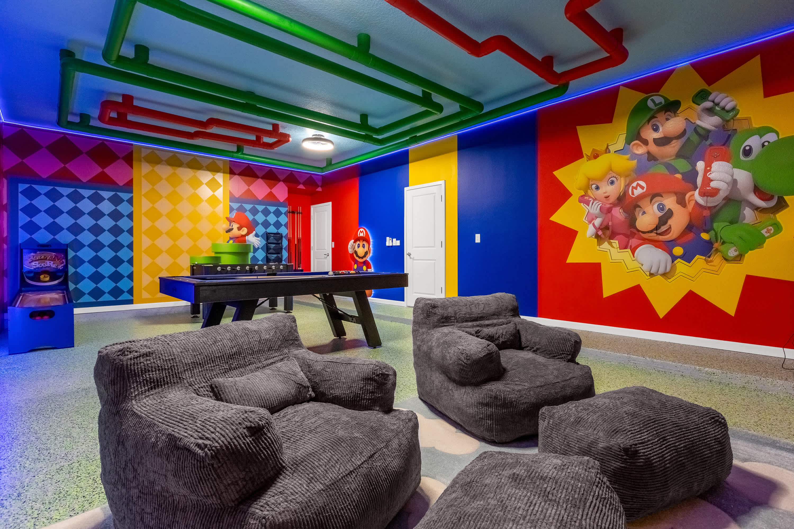 Expansive game room with a foosball table, two large grey bean bag chairs, and a vibrant Mario Bros. mural on a red and yellow wall, complemented by colorful overhead pipes and LED lighting.