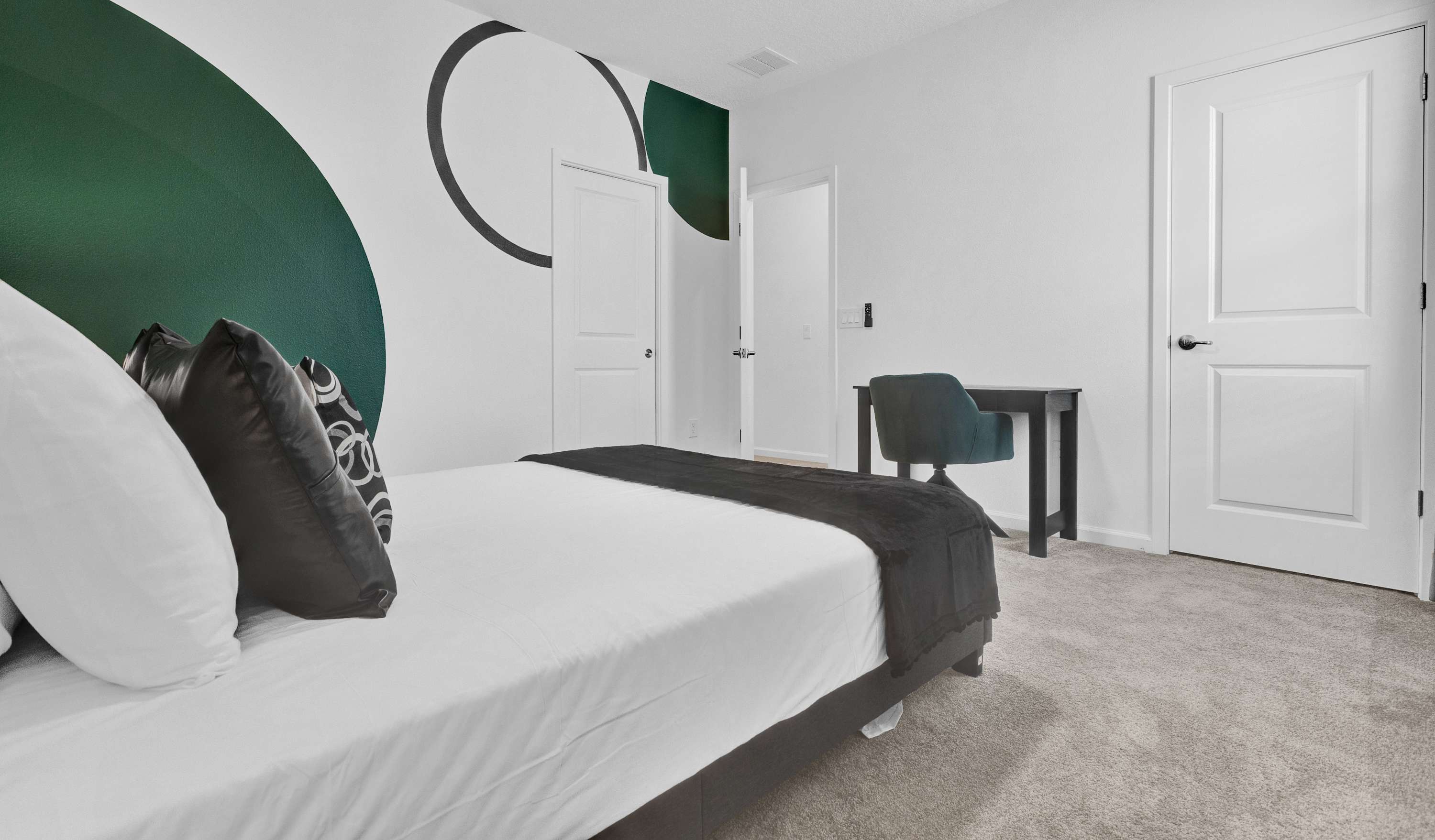 Another view of the contemporary bedroom, highlighting the green and black geometric wall mural and a dark desk with a teal chair in the corner. The room has gray carpet and white doors.