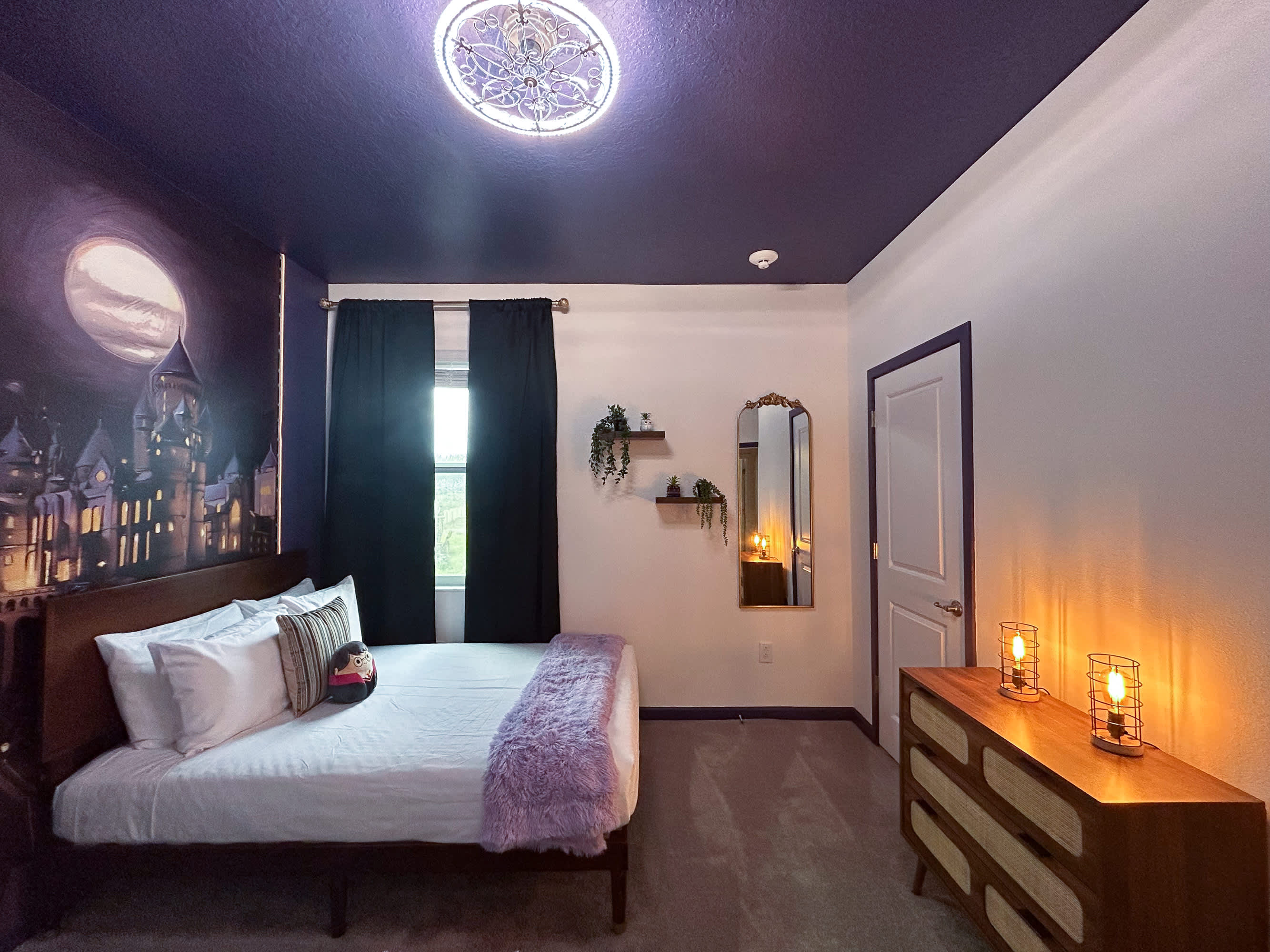 A wider view of the Harry Potter themed bedroom, showcasing the Hogwarts mural, dark purple ceiling, and a single twin bed with a purple throw. A window with dark curtains, floating shelves, and a decorative mirror are also visible.