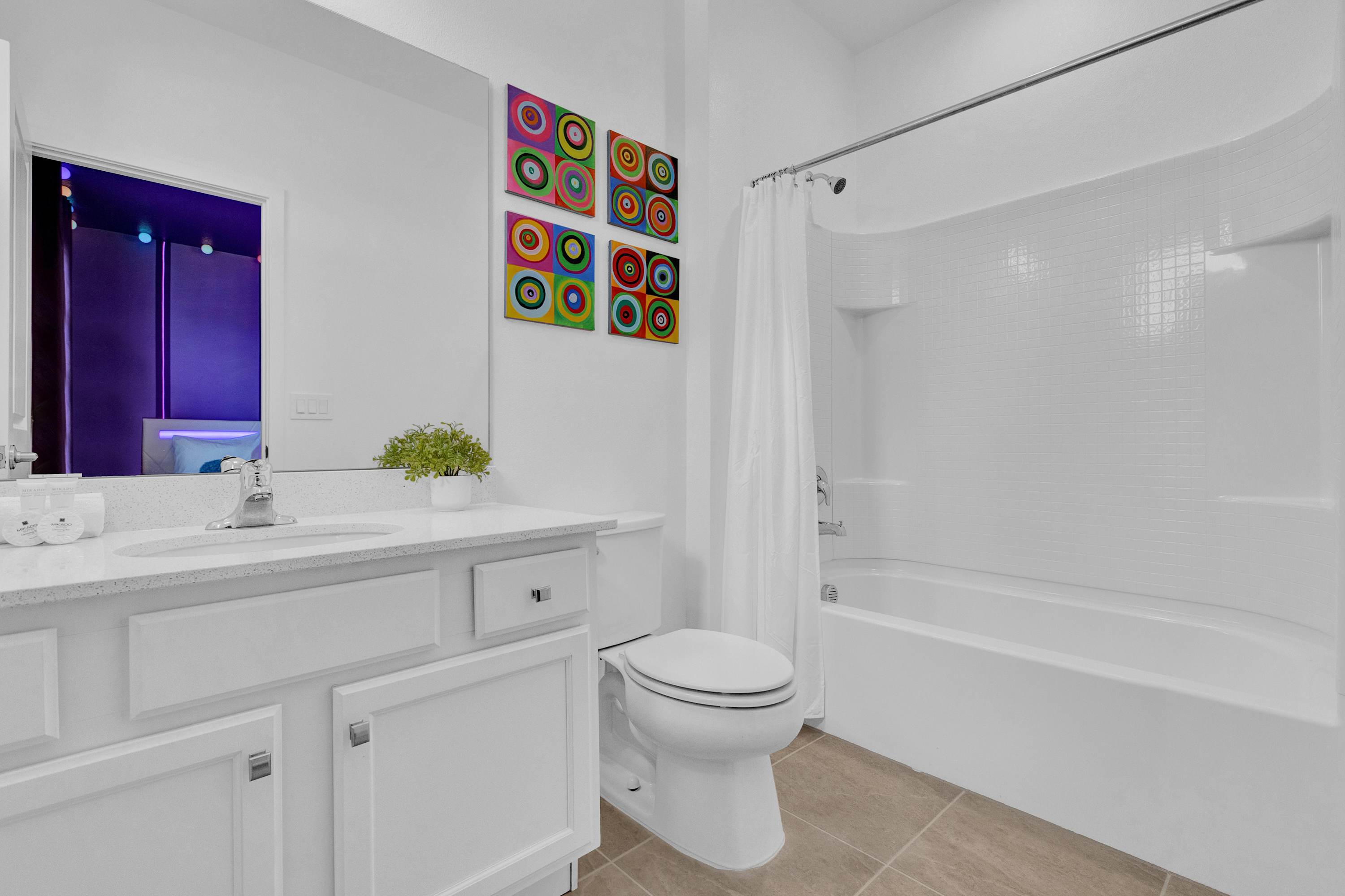 A clean, white bathroom featuring a single vanity with a white countertop and sink, a toilet, and a bathtub with a shower curtain, accented by colorful circular artwork on the wall.