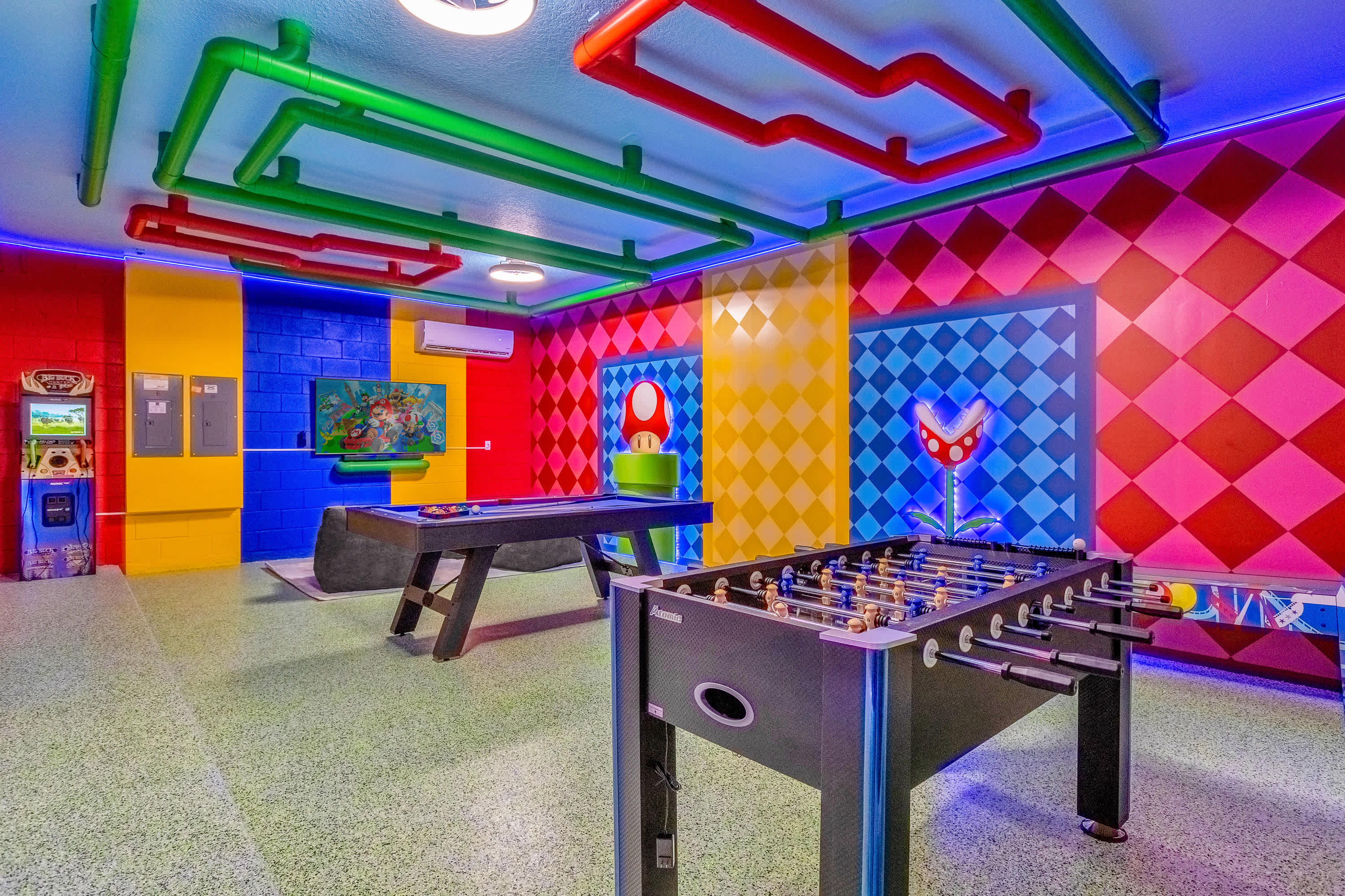 Vibrant, themed game room with a Super Mario Bros. design, featuring colorful walls, green and red pipes on the ceiling, a pool table, and a foosball table. Arcade machines are visible in the corner.
