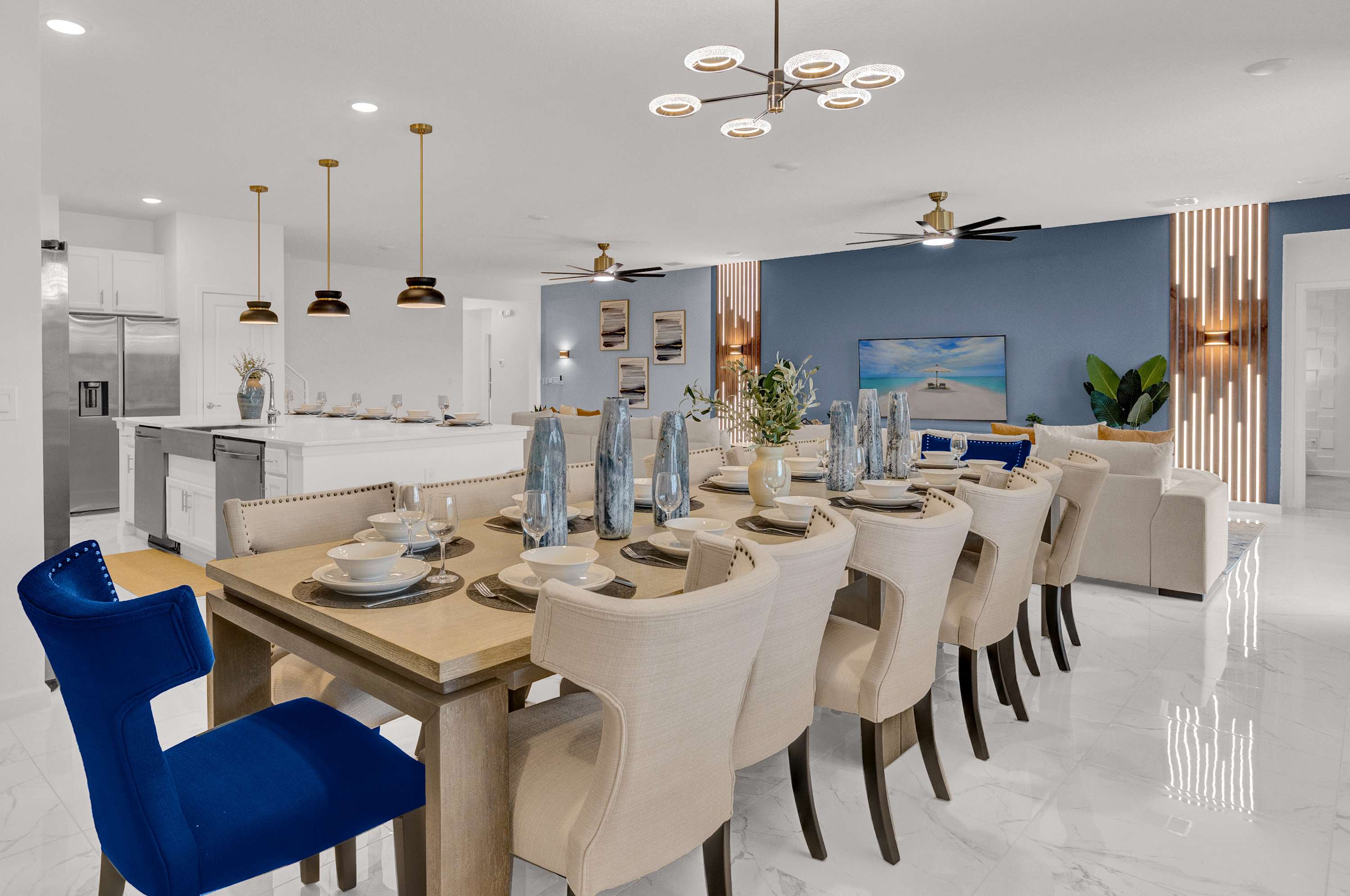 Elegant dining room with a long wooden table set for many guests, featuring light-colored upholstered chairs and two blue velvet chairs at the ends. A modern chandelier hangs above, with the kitchen and living area visible in the background.