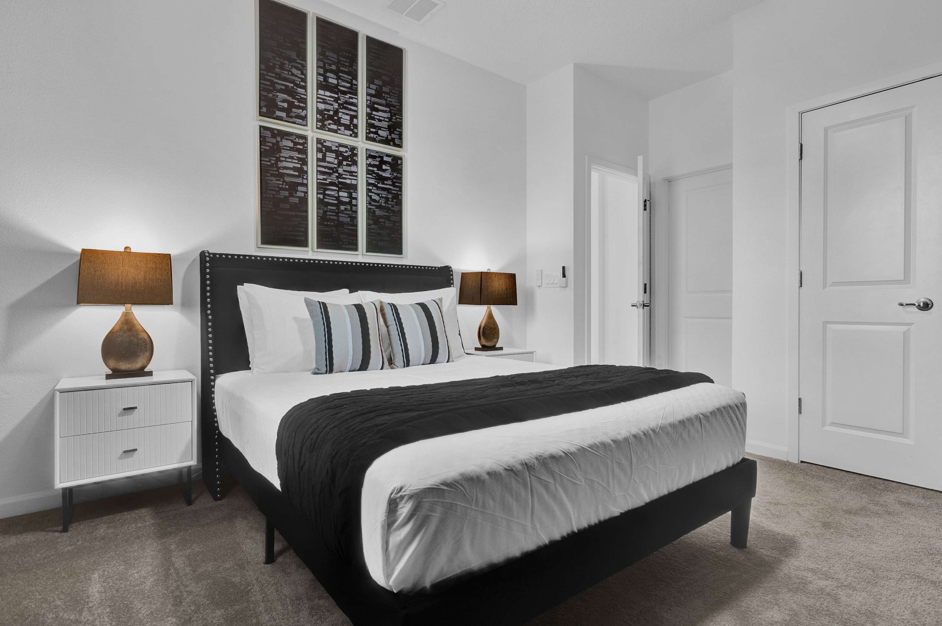 Modern bedroom with a black upholstered king bed, two white nightstands with bronze lamps, and a large multi-panel abstract artwork above the headboard. The room has gray carpet and white walls.