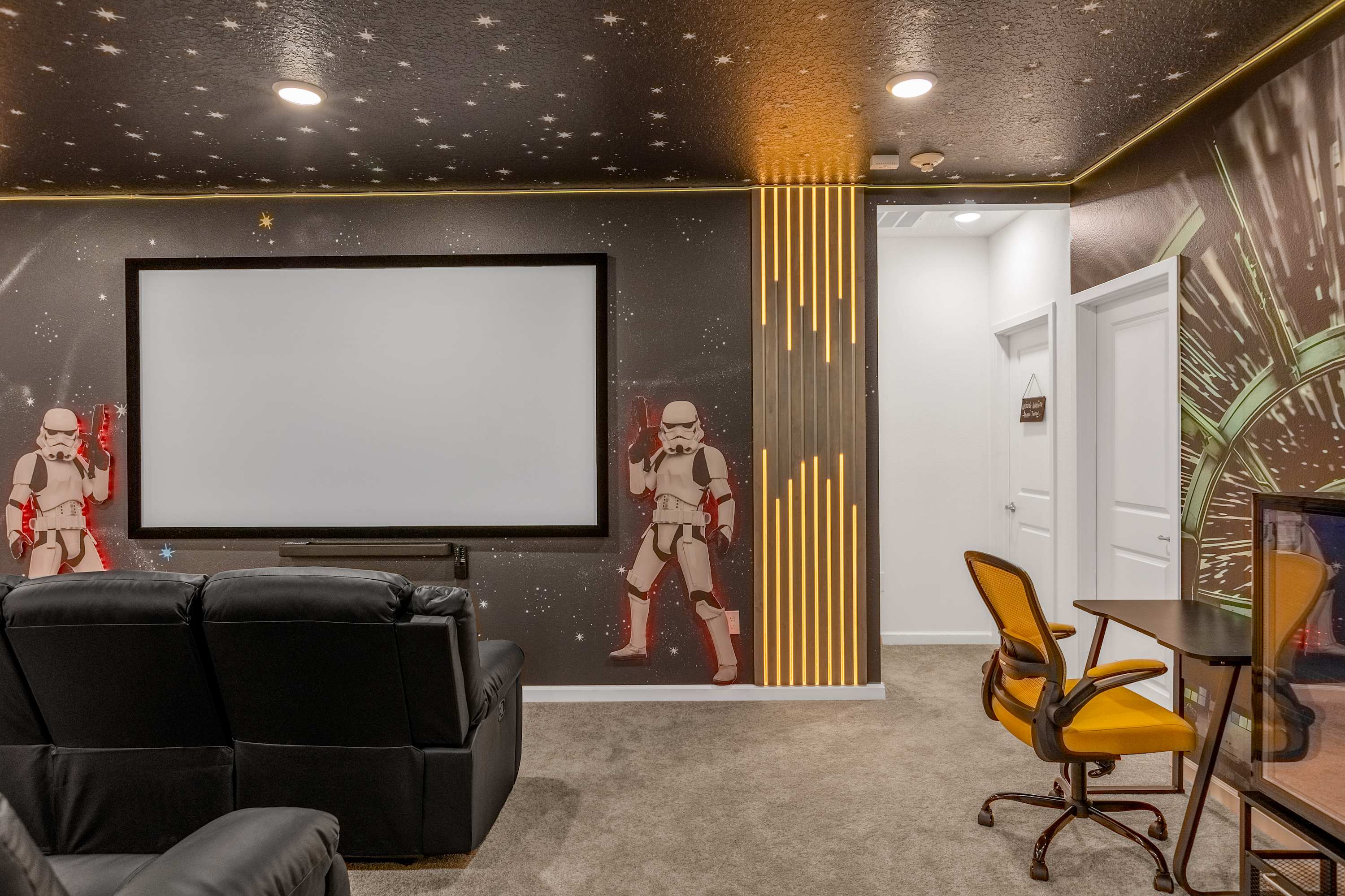 Detailed view of the media room showing a large projection screen flanked by Stormtrooper murals. A small desk with a yellow office chair is visible, along with decorative vertical light panels on the wall.