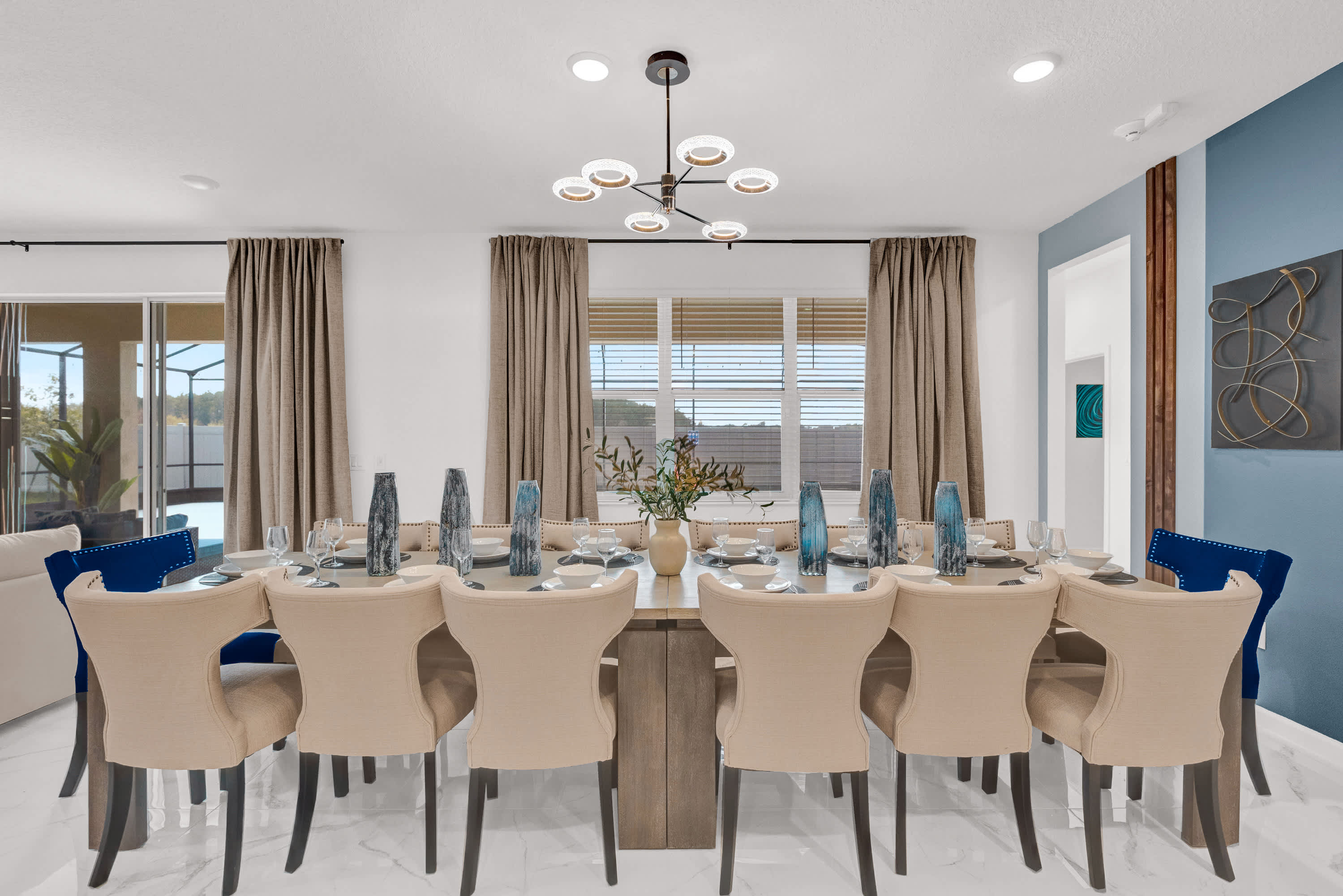 Formal dining area with a long wooden table, set with place settings and decorative vases, flanked by light-colored upholstered chairs and two blue velvet chairs. Large windows with blinds and curtains are visible in the background.