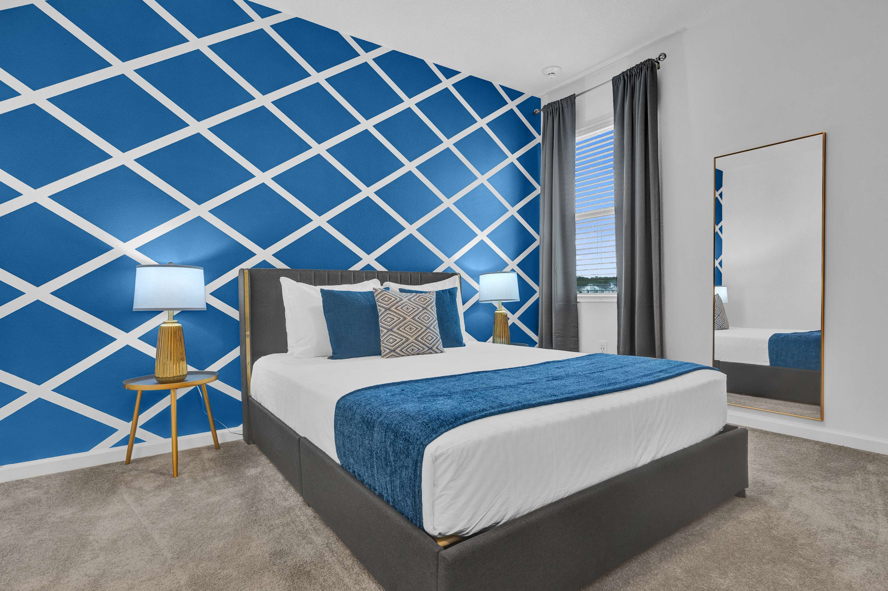 Vibrant bedroom featuring a bold blue accent wall with a white geometric pattern, a king bed with a gray upholstered frame, and two gold-legged nightstands with modern lamps. A full-length mirror leans against the wall.