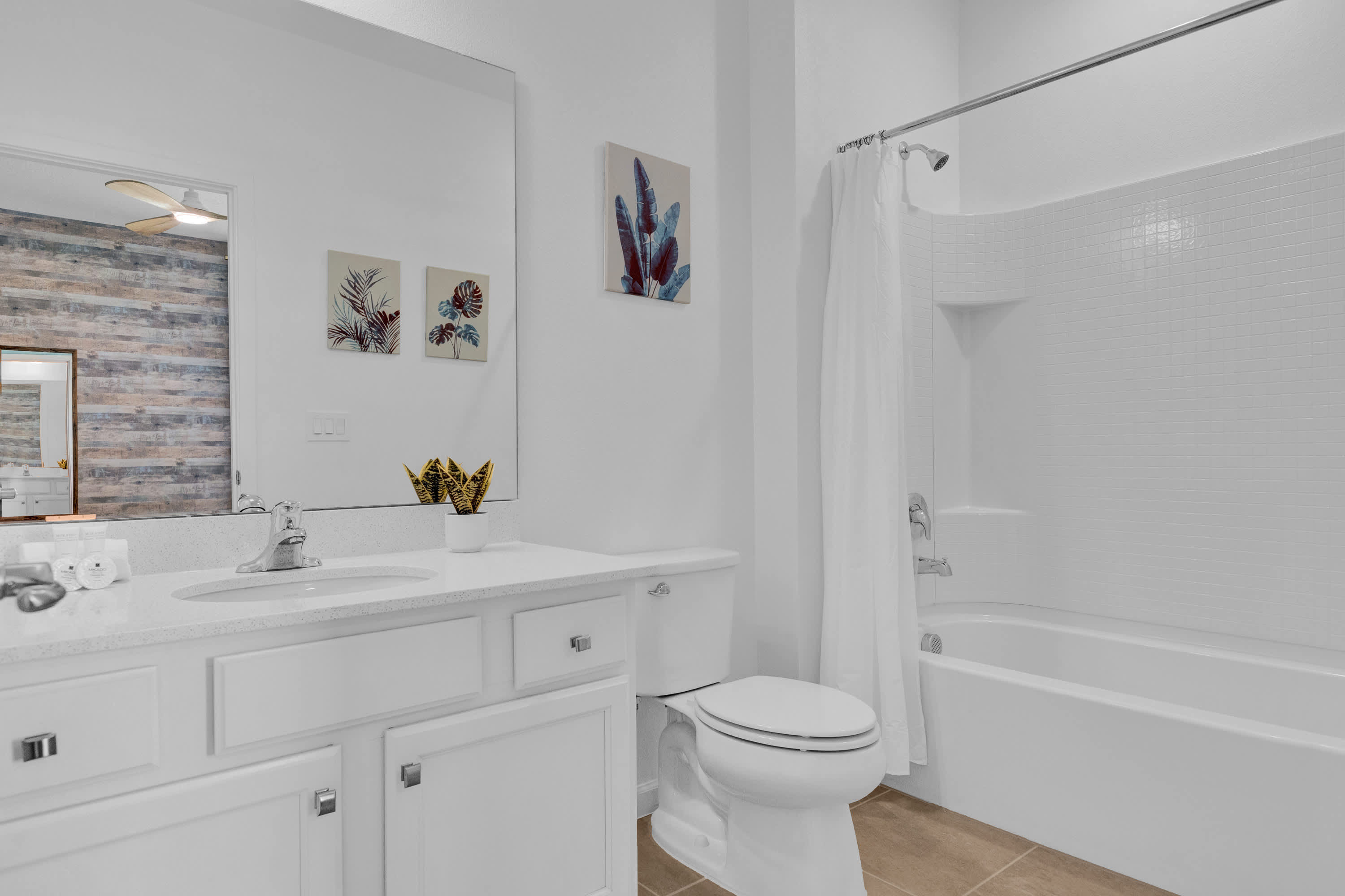 A clean white bathroom featuring a single vanity with a white countertop, a large mirror, and a toilet. A white tiled tub and shower combo with a shower curtain is visible on the right.