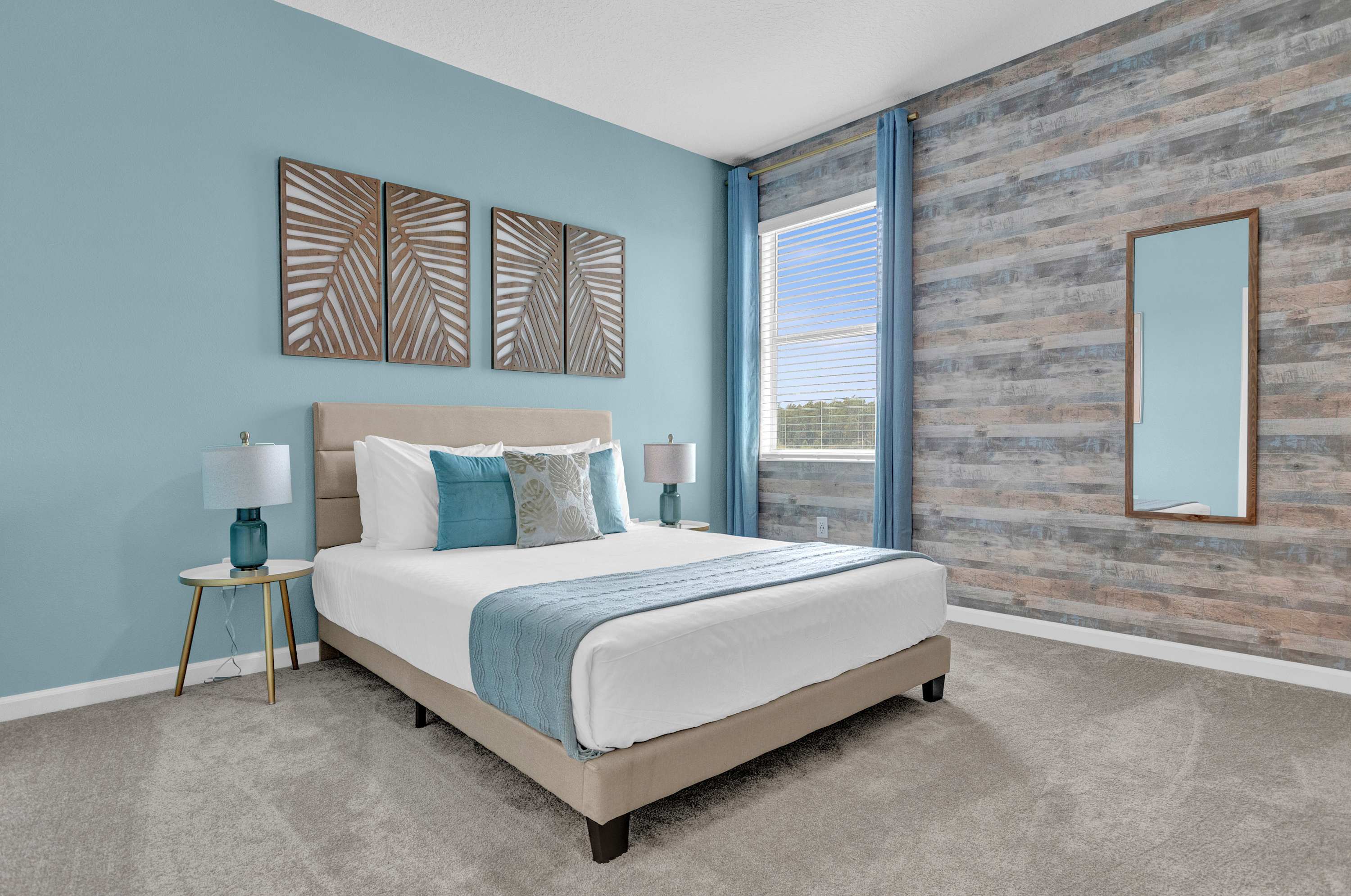 A bright bedroom with light blue walls, a beige upholstered bed, and a feature wall with horizontal wood paneling. Two wooden art pieces hang above the bed, and a tall mirror leans against the paneled wall.