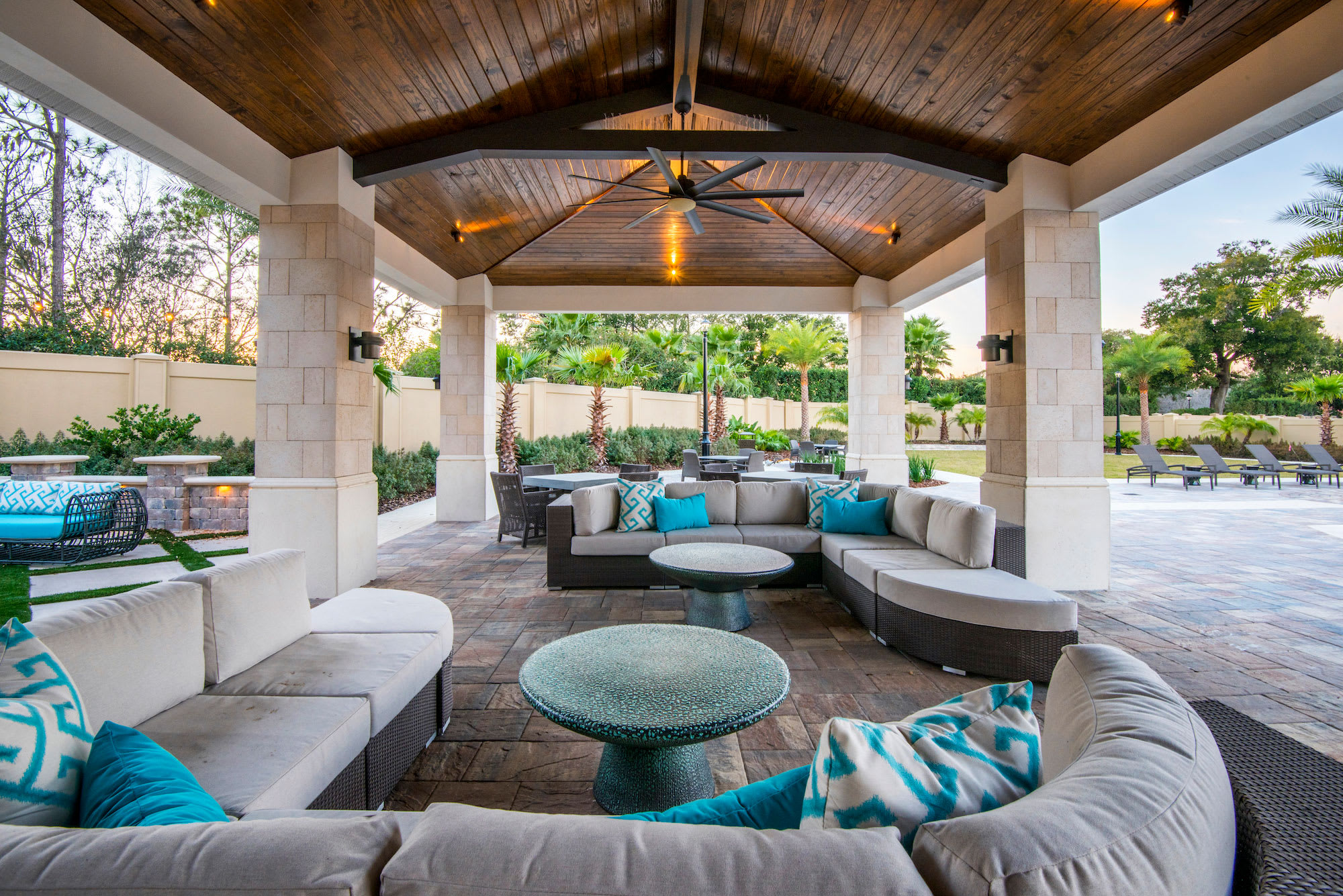 A spacious covered outdoor patio features comfortable L-shaped sectional sofas with teal accent pillows, arranged around round coffee tables on a patterned stone floor. The area is illuminated by a wooden plank ceiling with a fan, overlooking a landscaped yard.