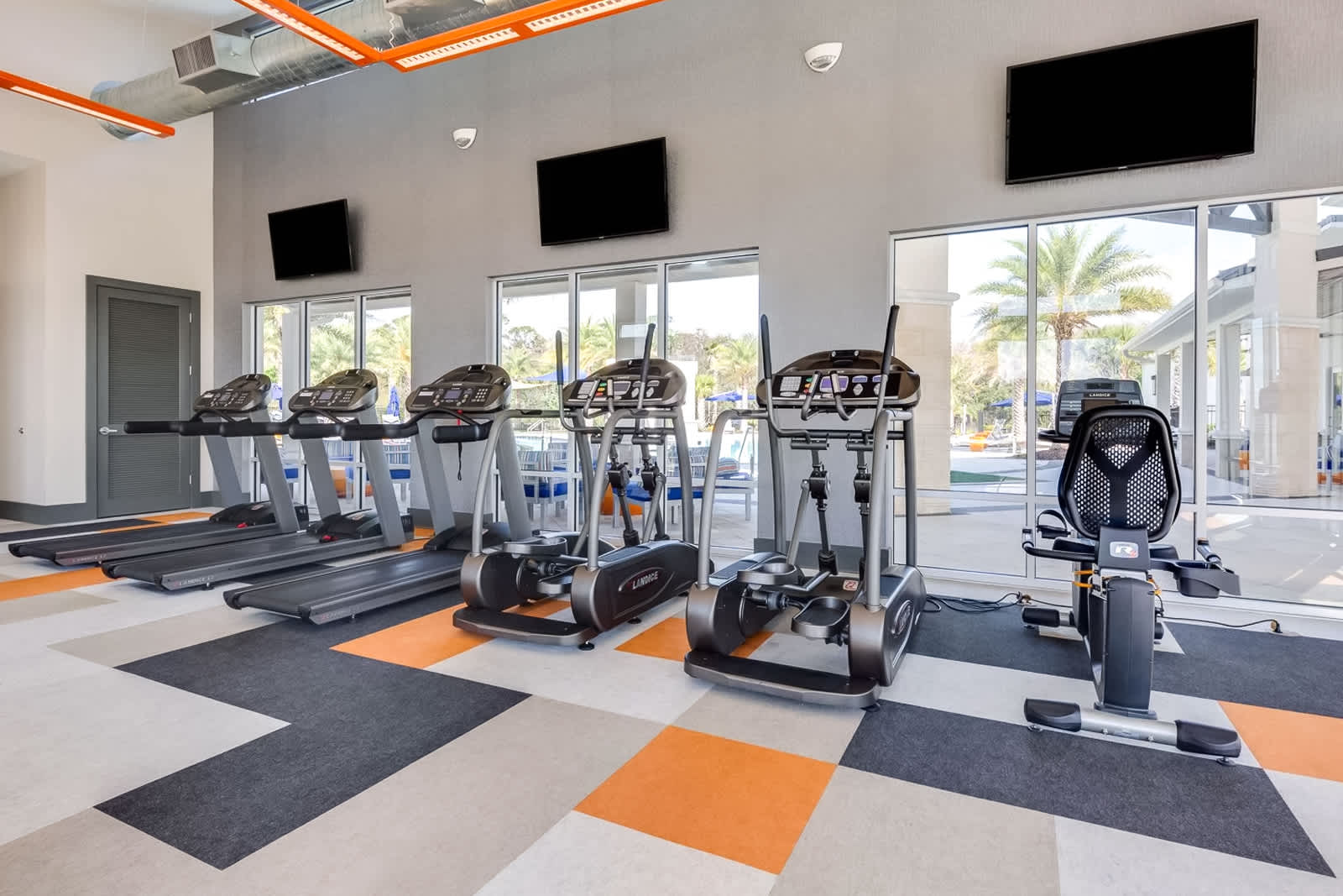 A modern fitness center features a row of treadmills, ellipticals, and stationary bikes, all facing large windows that offer views of an outdoor pool area. The room has a colorful geometric carpet and wall-mounted TVs.