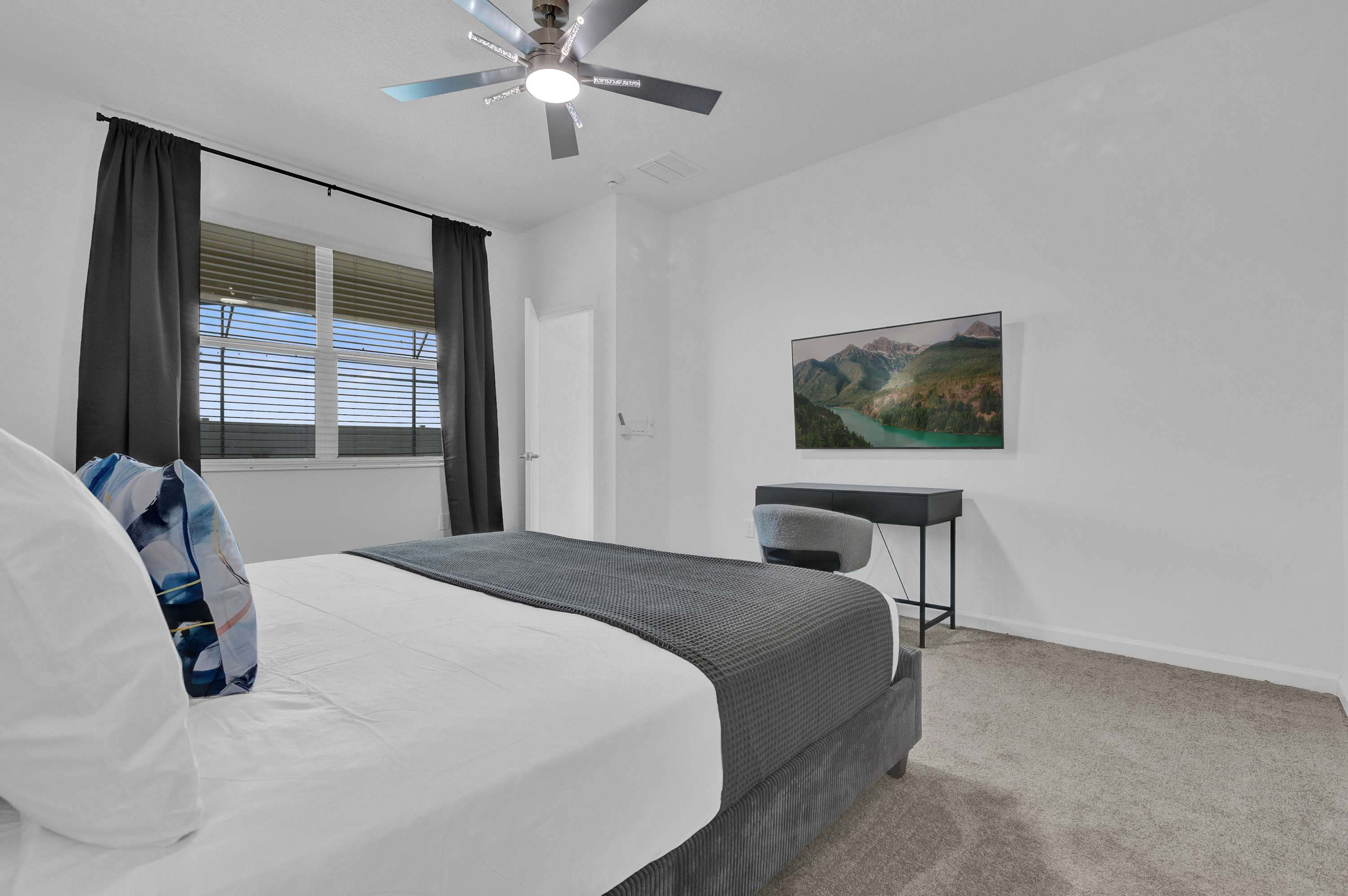 View of bedroom-2 showing a wall-mounted flat-screen TV opposite the bed, a sleek black desk with a chair, and a window with black curtains, providing a functional workspace.